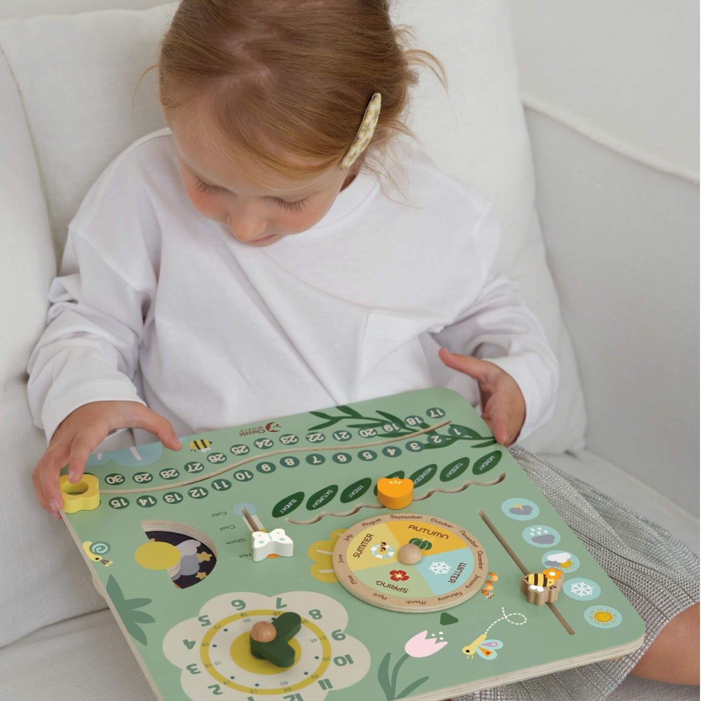 Young child sitting on a sofa playing with a pastel green wooden learning board, moving the dials and sliders to explore days, months, and weather.