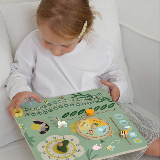 Young child sitting on a sofa playing with a pastel green wooden learning board, moving the dials and sliders to explore days, months, and weather.