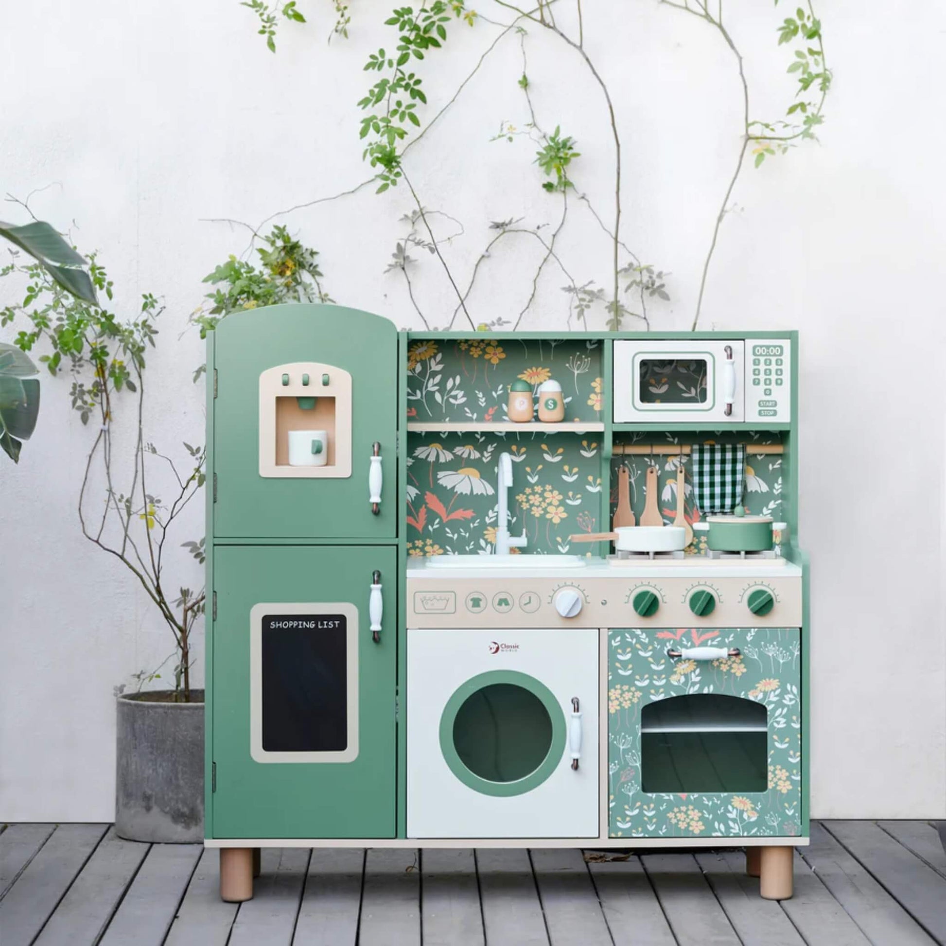 A full play kitchen set is displayed outdoors against a white wall, with potted plants and trailing vines creating a calm, natural setting around the toy.