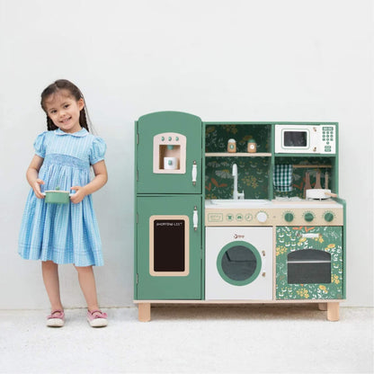 A smiling young girl in a blue dress stands next to a toy kitchen set, holding a toy pot and looking proud of her pretend cooking setup.