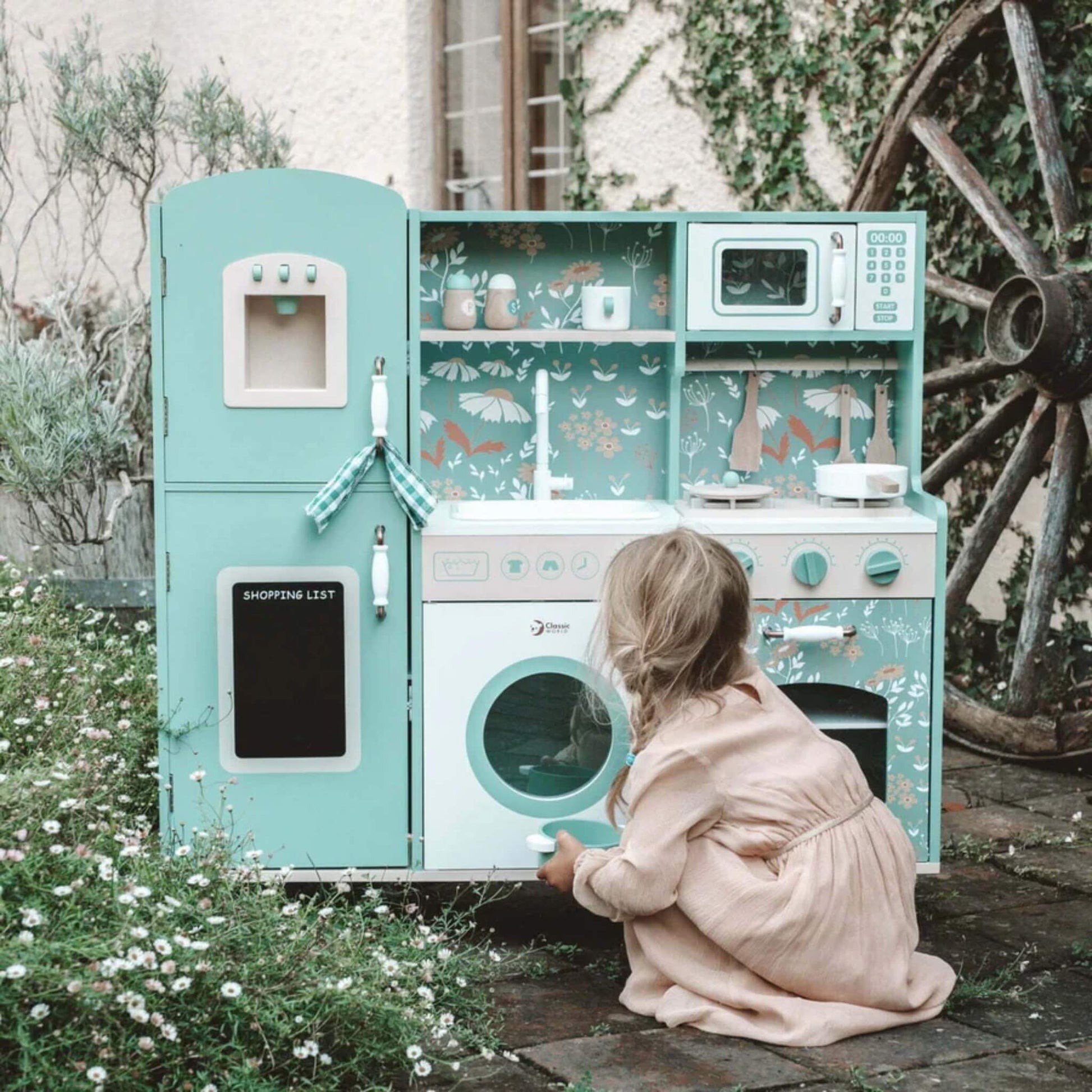 A child in a peach-coloured dress crouches on a stone patio, opening the toy washing machine door on a green wooden play kitchen surrounded by wildflowers.