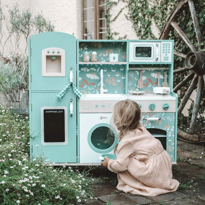 A child in a peach-coloured dress crouches on a stone patio, opening the toy washing machine door on a green wooden play kitchen surrounded by wildflowers.