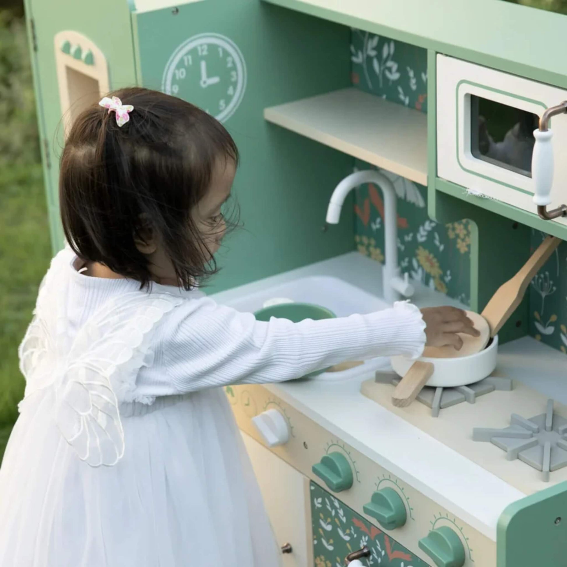 A child dressed in a white fairy-style outfit stirs a wooden pot on the hob of a green and cream play kitchen in a grassy outdoor setting.