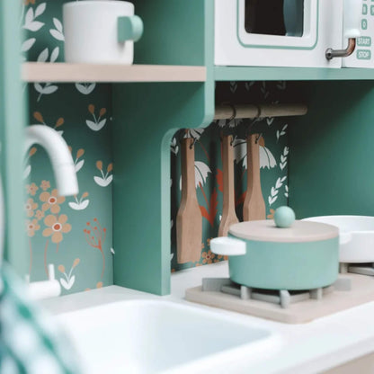 A close-up of the kitchen’s countertop shows hanging utensils, a toy pot on the stove, a sink, and a shelf with a cup, all set against a floral green backdrop.