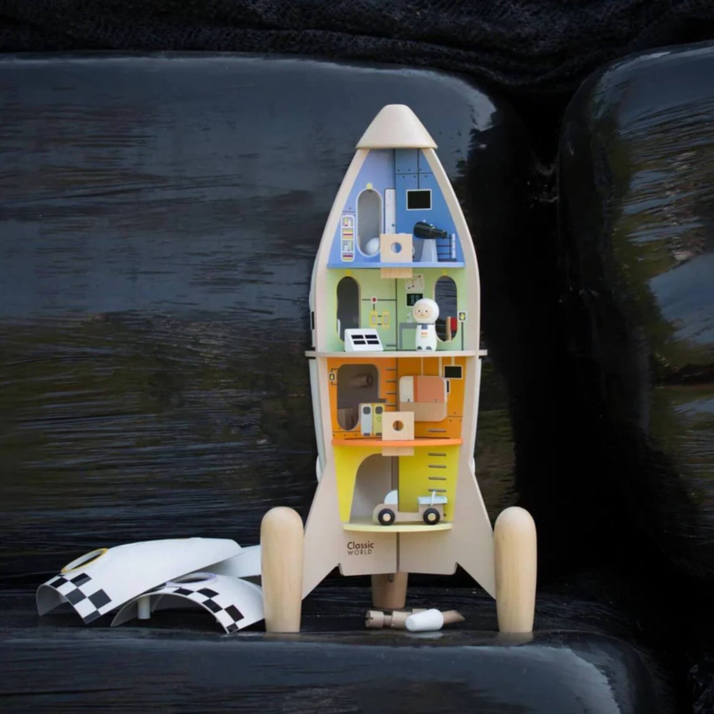 A vertical view of the rocket playset placed on dark outdoor decking, with the interior fully visible and play accessories including astronauts and a scooter scattered in front.
