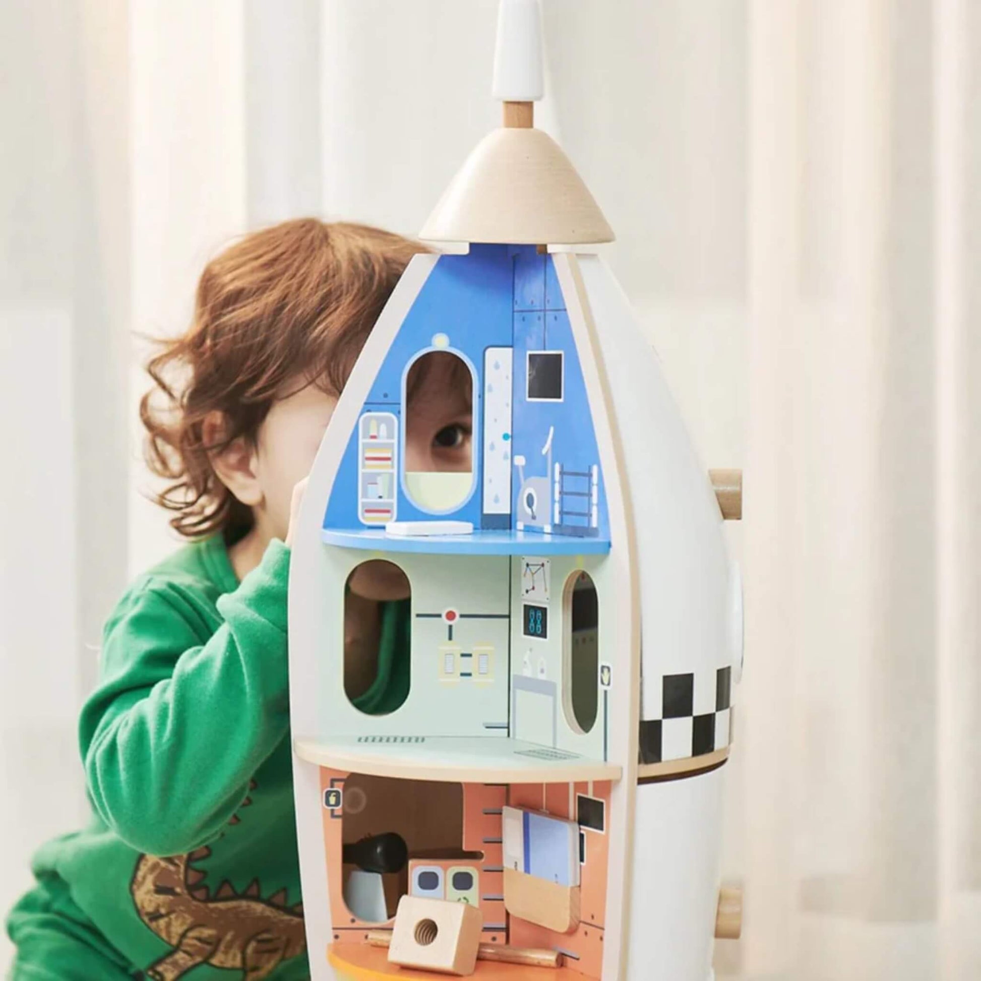 A young child wearing a green top peers into the top level of a wooden rocket playset, engaging with its colourful rooms and space-themed printed details.