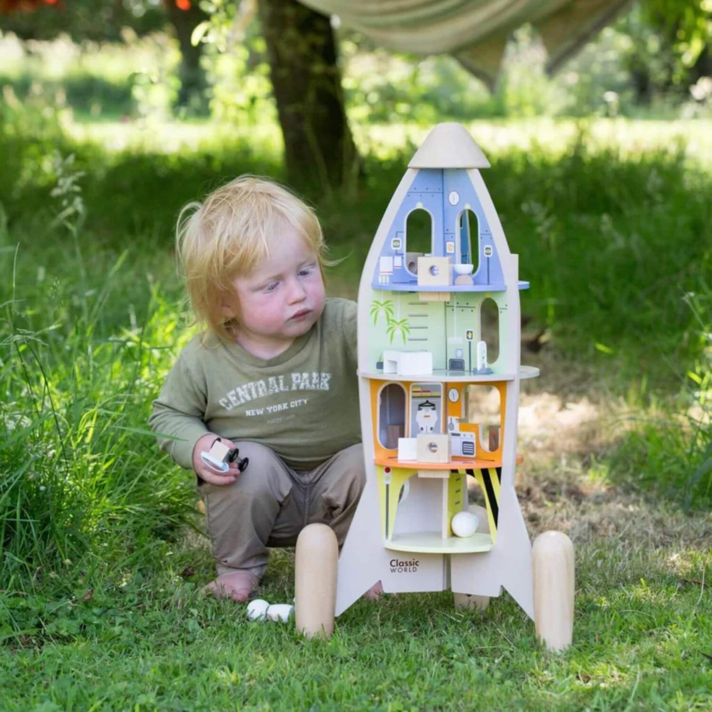 A toddler sits in the grass beside the opened rocket playset, exploring its detailed levels and small accessories in an outdoor garden setting.