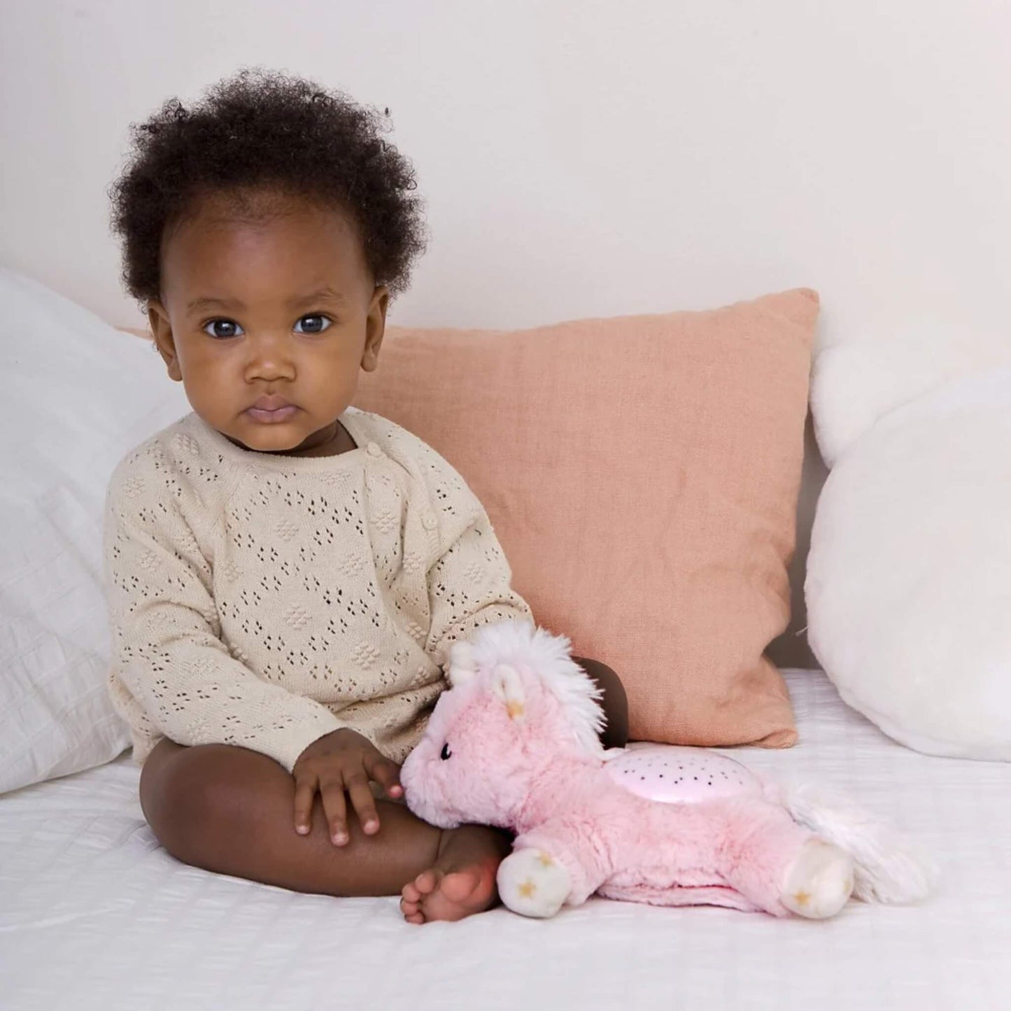 A baby sitting upright on a bed beside a pink unicorn plush night-light, with soft cushions in the background creating a cosy setting.
