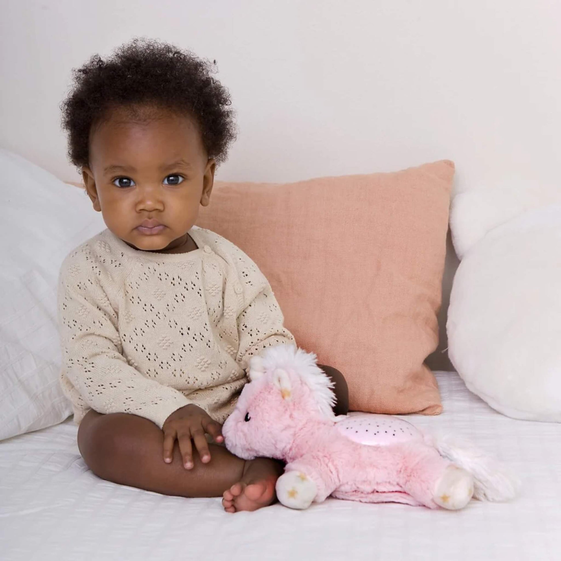 A baby sitting upright on a bed beside a pink unicorn plush night-light, with soft cushions in the background creating a cosy setting.
