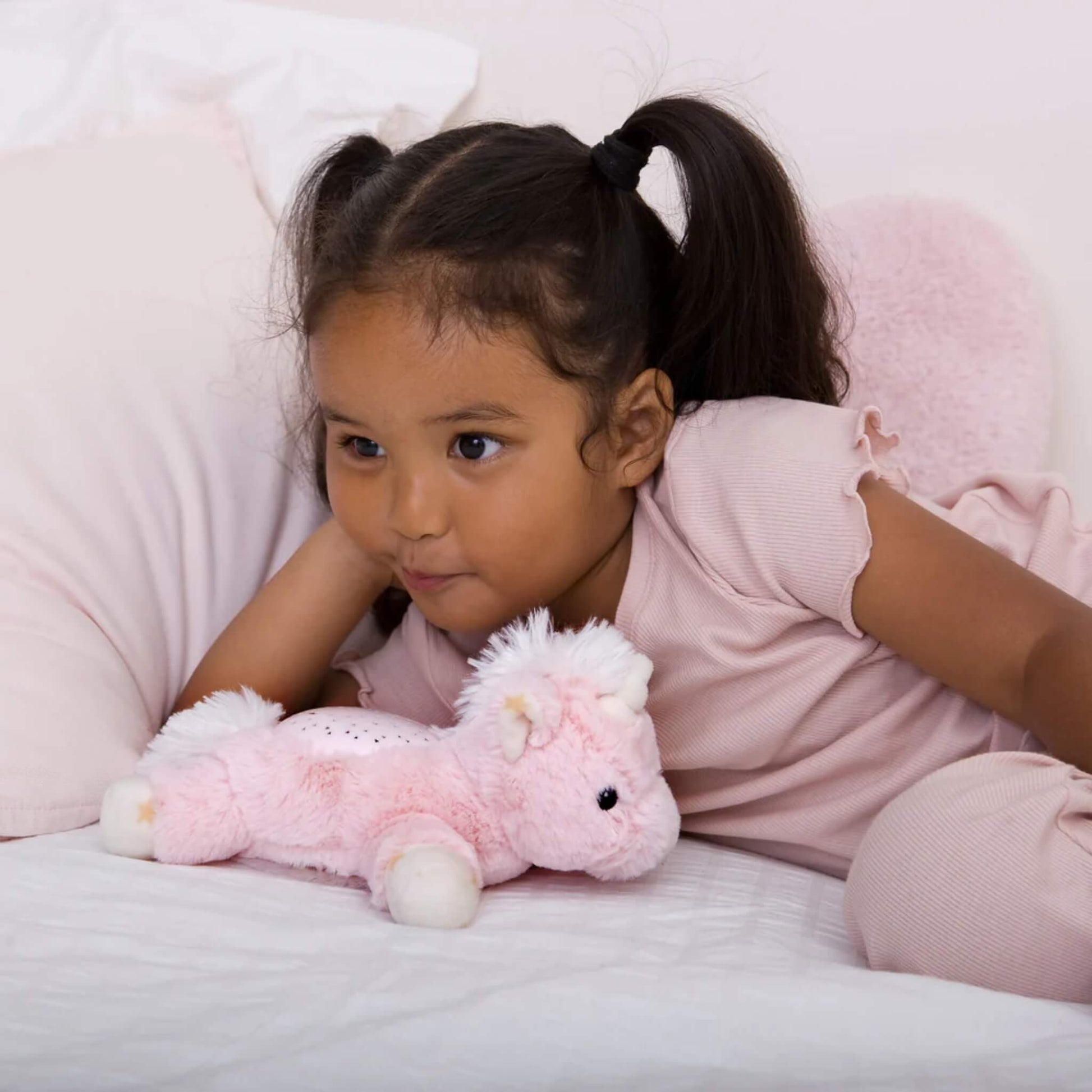 A child lying on a bed with a pink unicorn plush night-light resting beside them, looking comfortable and relaxed.