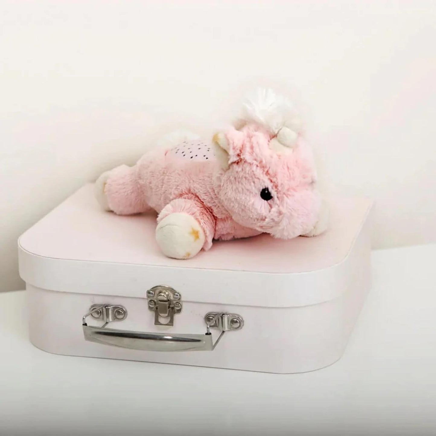 The unicorn plush night-light placed on top of a small pastel pink suitcase with silver clasp, showing its compact and portable size.