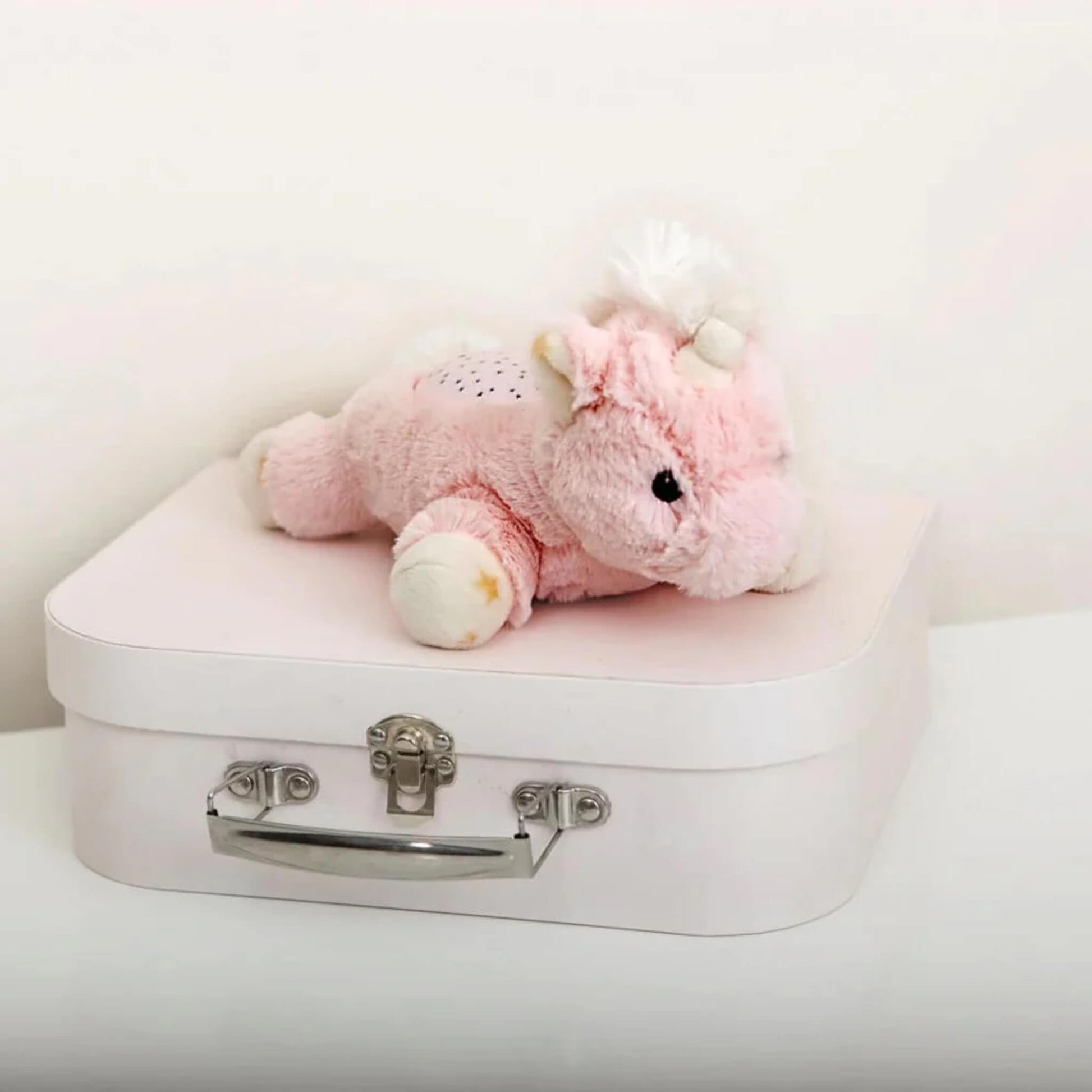 The unicorn plush night-light placed on top of a small pastel pink suitcase with silver clasp, showing its compact and portable size.
