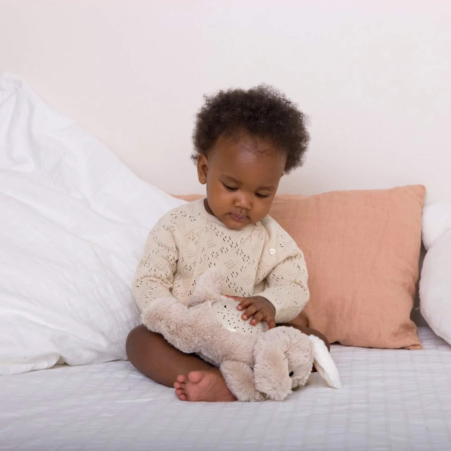 Baby sitting on a bed wearing a cream knitted outfit, gently holding a beige plush puppy night-light toy across their lap.