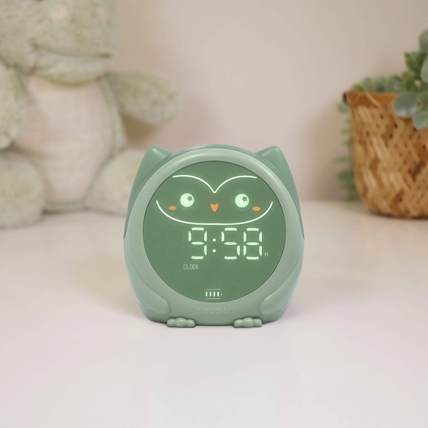 A green owl-shaped sleep trainer  clock displaying the time on a bedside table.