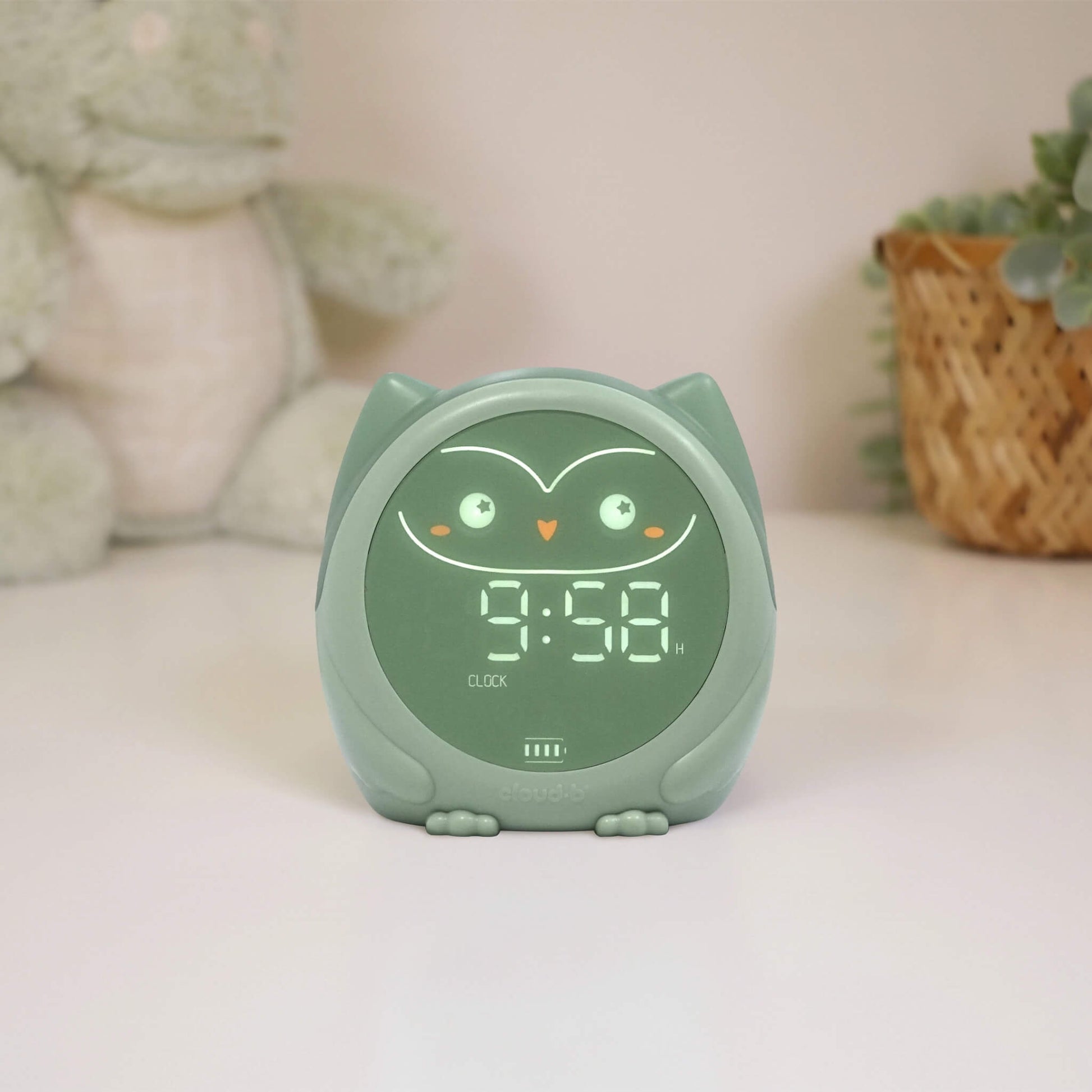 A green owl-shaped sleep trainer  clock displaying the time on a bedside table.