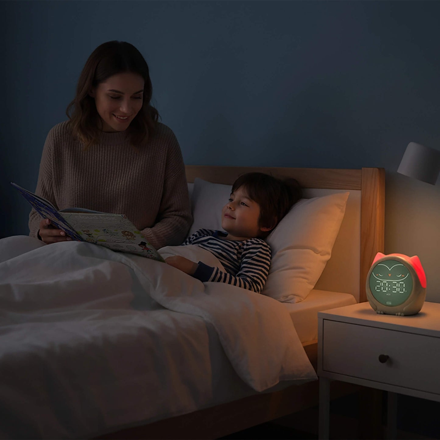 A mother and child are reading in bed at night, with a smart owl alarm clock glowing on the bedside table. The alarm clock has a green owl face and a pinkish-red glowing eye.