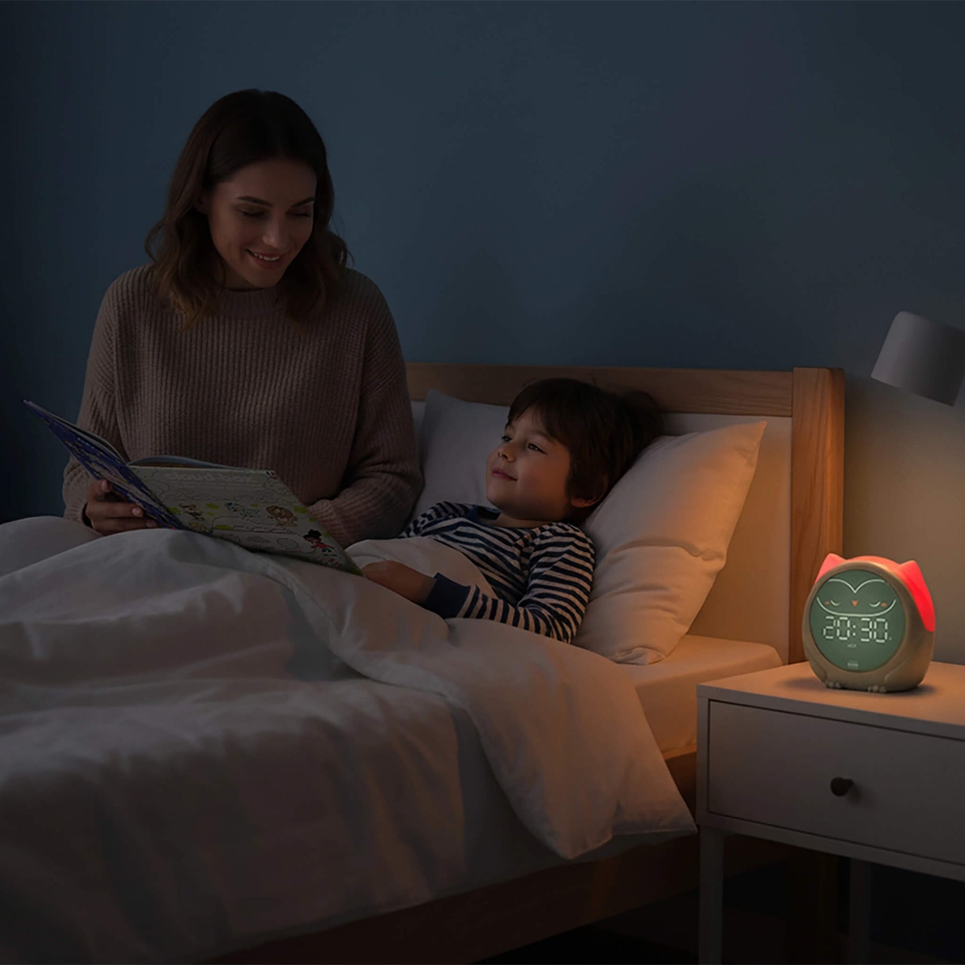 A mother and child are reading in bed at night, with a smart owl alarm clock glowing on the bedside table. The alarm clock has a green owl face and a pinkish-red glowing eye.