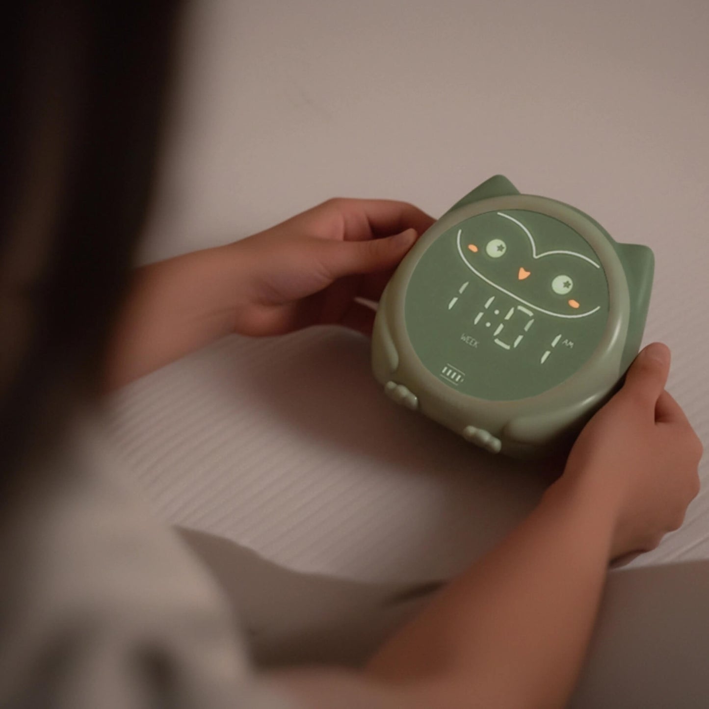 A hand adjusting a green owl sleep trainer clock while it rests on a bed.
