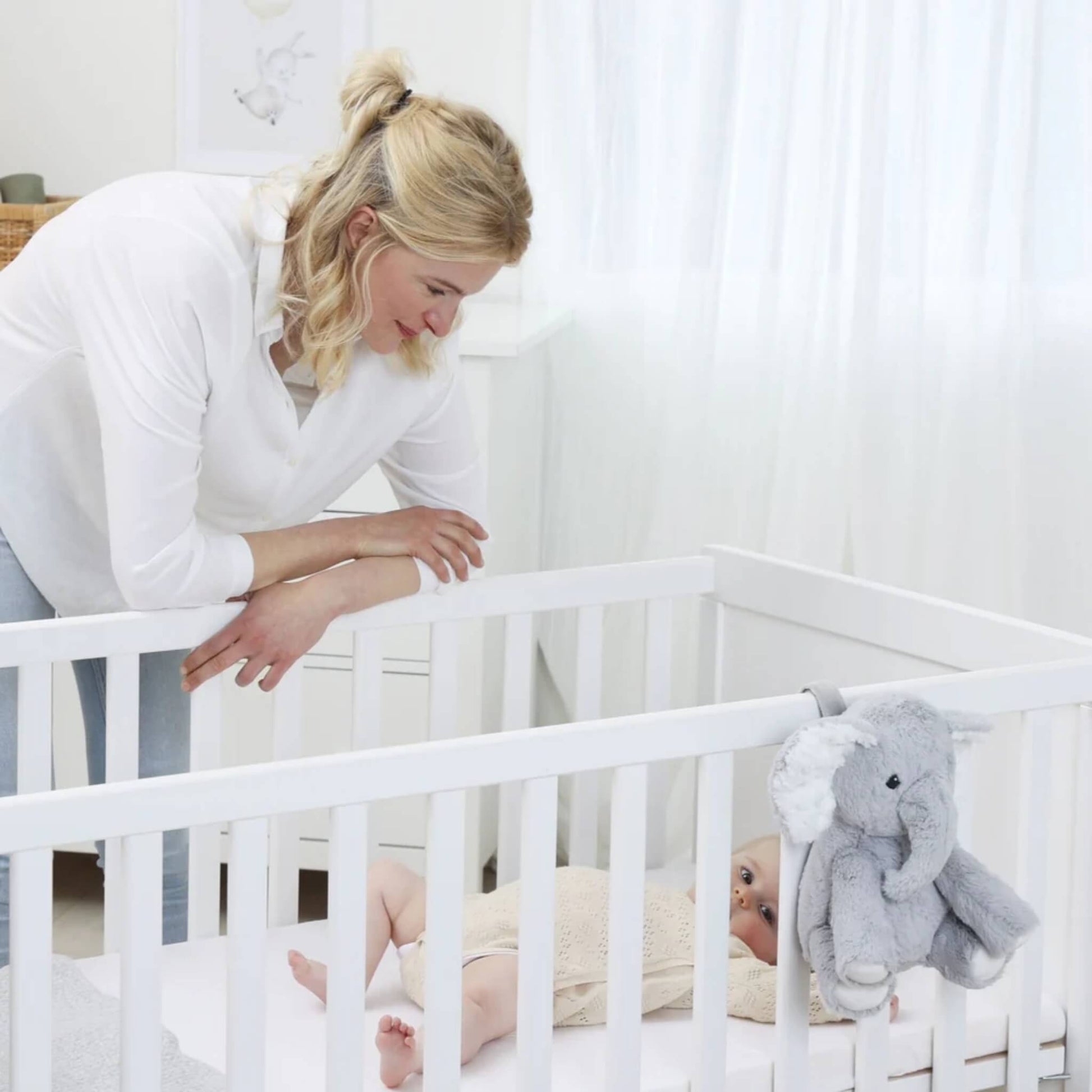 Mother leaning over a white cot where a baby lies on their back, with a plush grey elephant sound soother attached securely to the cot rail.