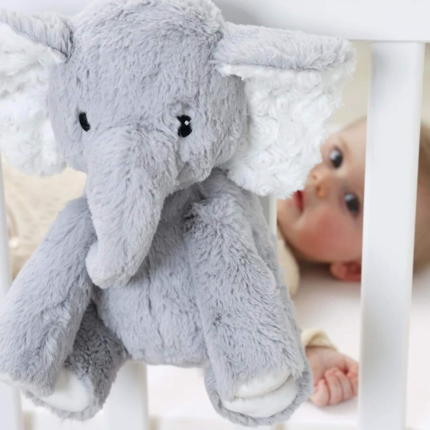 Close-up of plush elephant sound soother hanging on white cot rail, with a baby lying inside the cot looking directly at the toy.