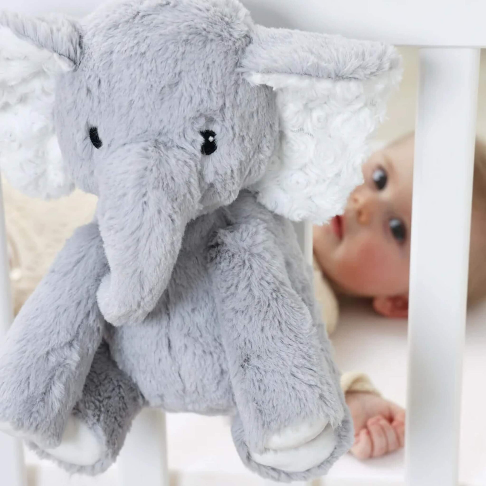 Close-up of plush elephant sound soother hanging on white cot rail, with a baby lying inside the cot looking directly at the toy.