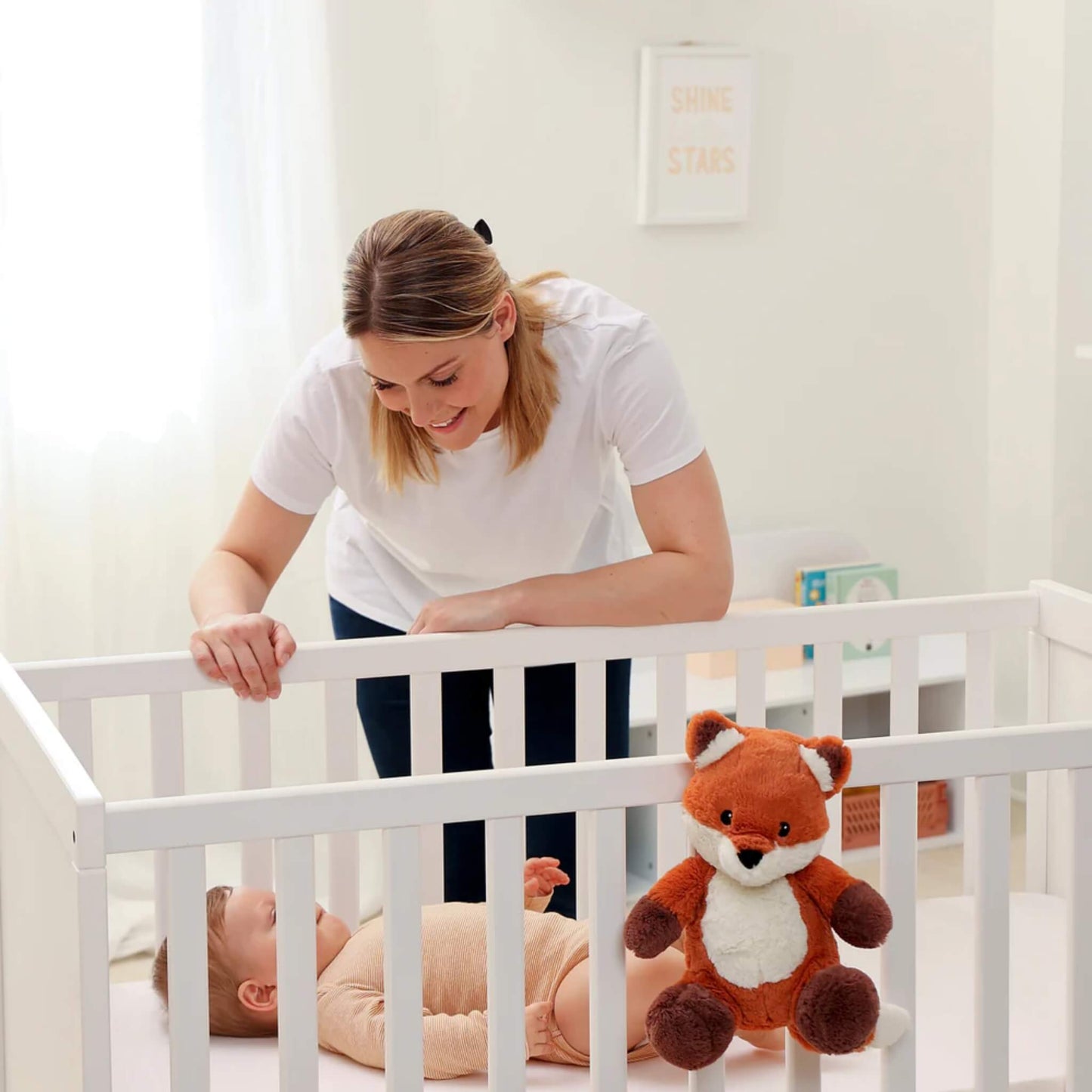 Mother leaning over a cot smiling at her baby, with a plush fox sound soother strapped to the cot rail beside the child.