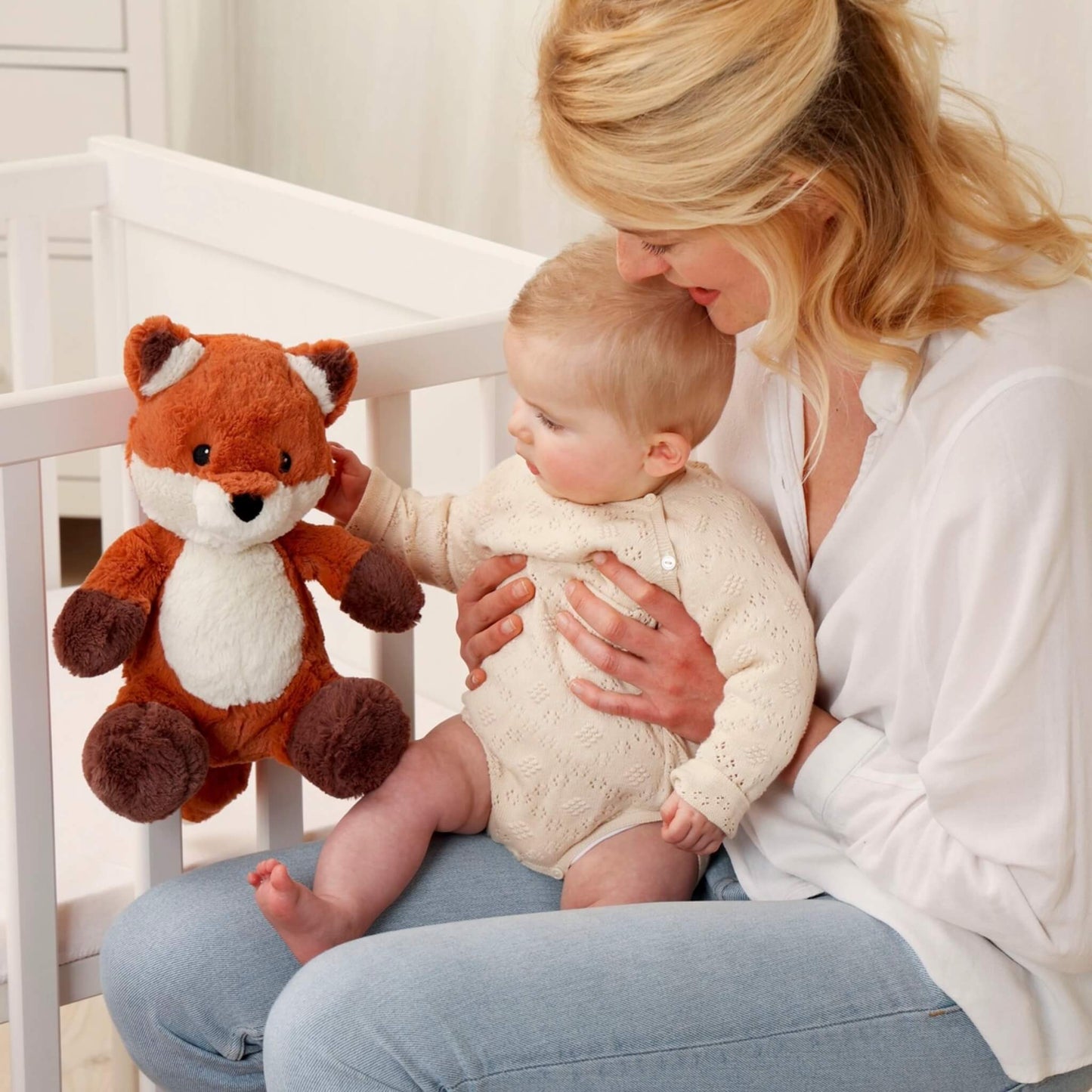 Mother holding her baby near a cot while the baby reaches out to touch a plush fox sound soother attached to the rail.