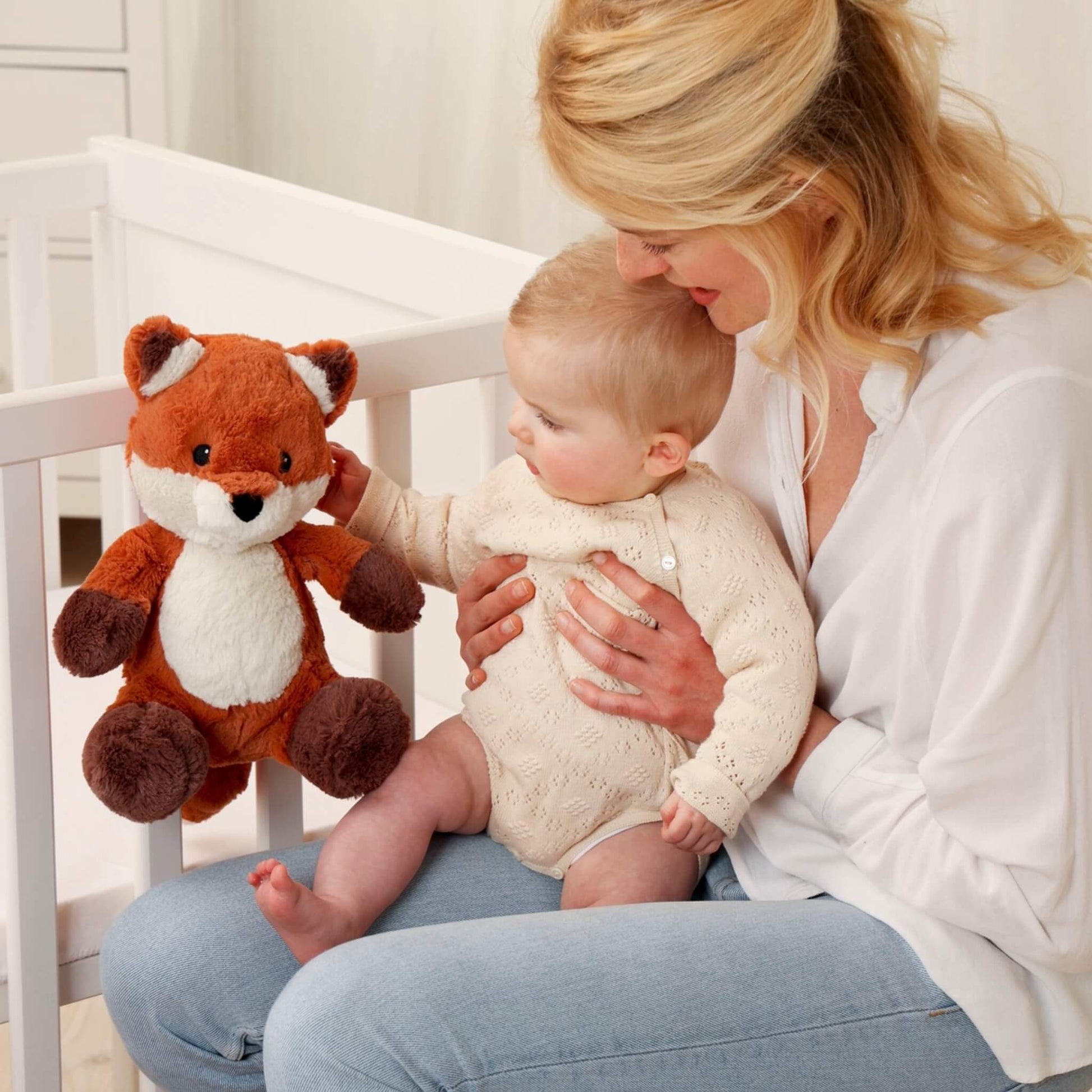 Mother holding her baby near a cot while the baby reaches out to touch a plush fox sound soother attached to the rail.