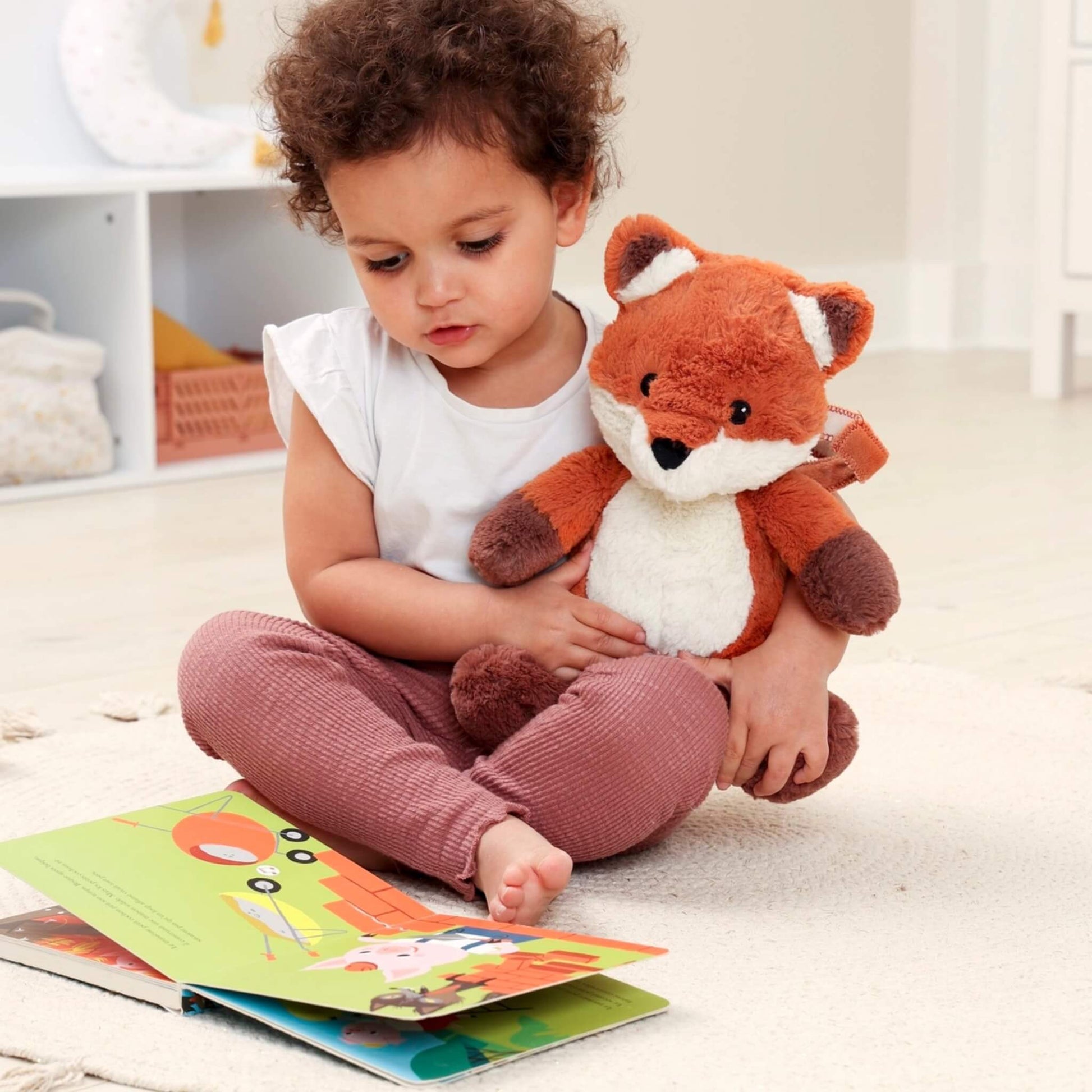 Young child sitting on the floor reading a picture book while holding a plush fox sound soother on their lap.