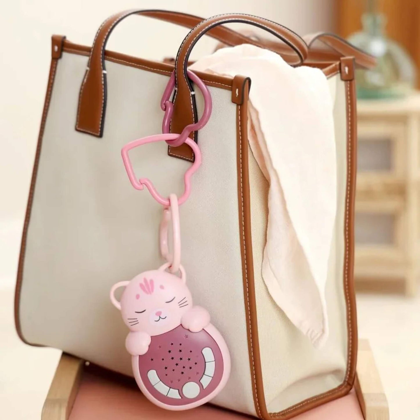 A pink cat-shaped portable baby white noise sleep aid clipped to the handle of a cream and brown tote bag with a heart-shaped link, showing how it can be carried when travelling.