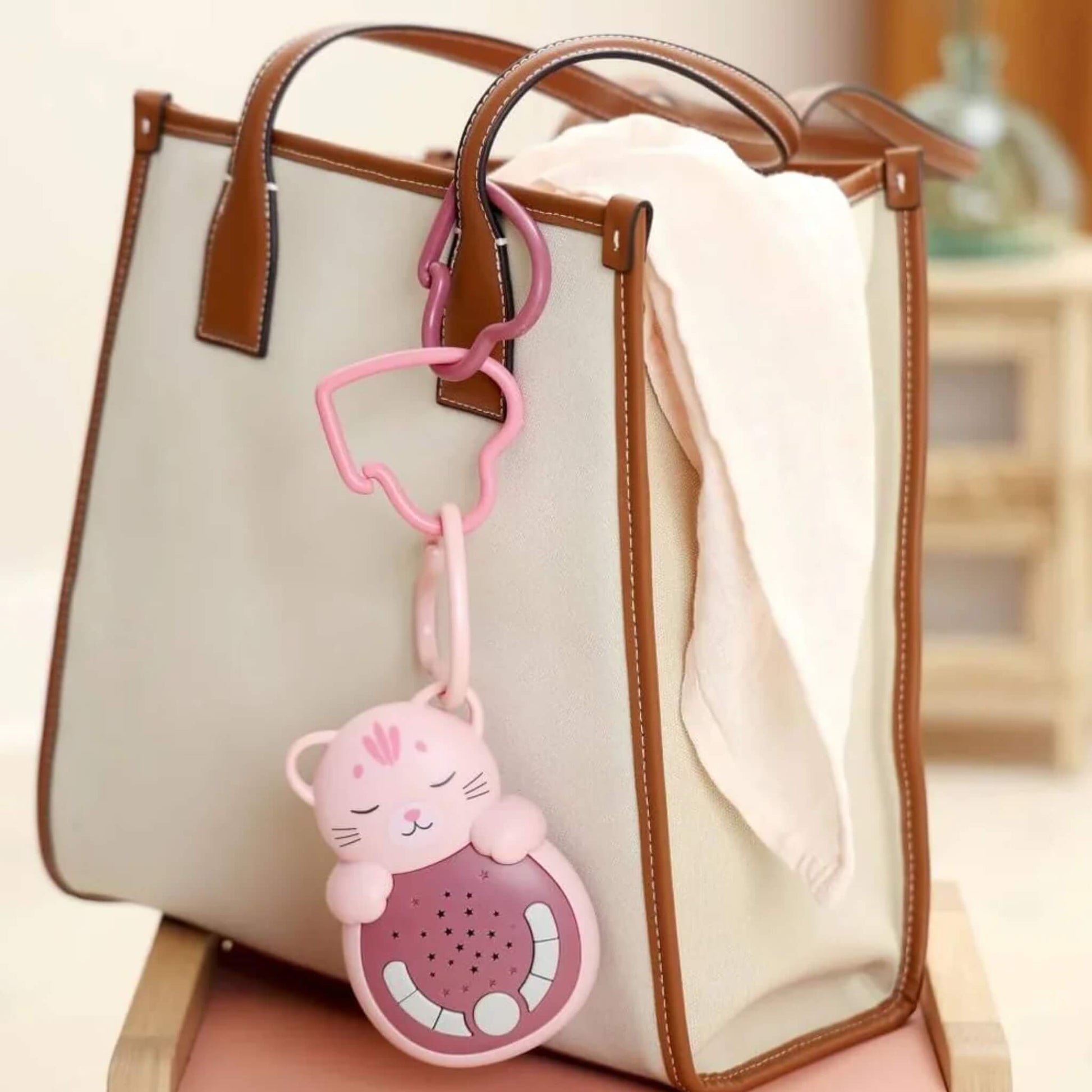 A pink cat-shaped portable baby white noise sleep aid clipped to the handle of a cream and brown tote bag with a heart-shaped link, showing how it can be carried when travelling.