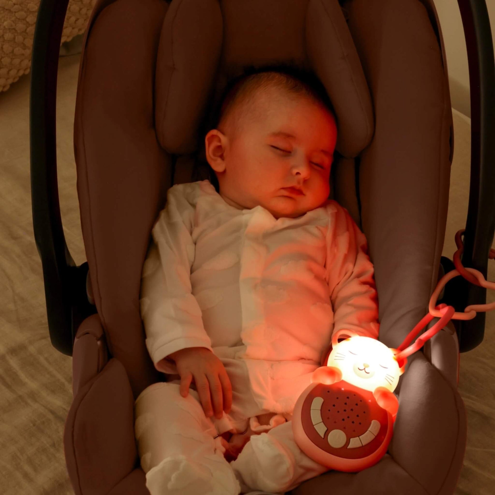A sleeping baby in a car seat illuminated by a gentle red glow from a clipped-on cat-shaped night-light, offering comfort during journeys.