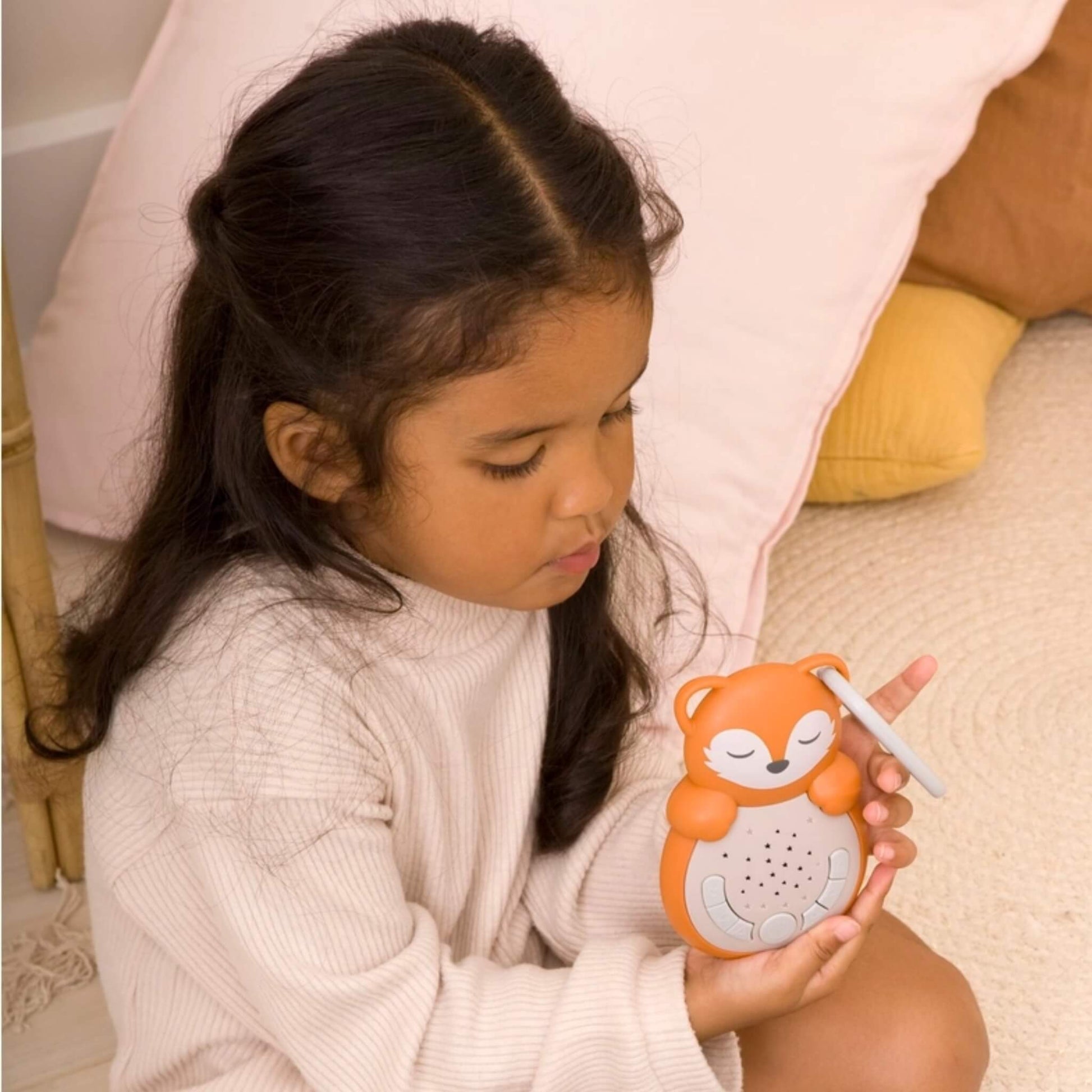 A young child in a cream jumper sitting on the floor, holding an orange fox-shaped sleep aid with a grey clip, looking down at it thoughtfully.