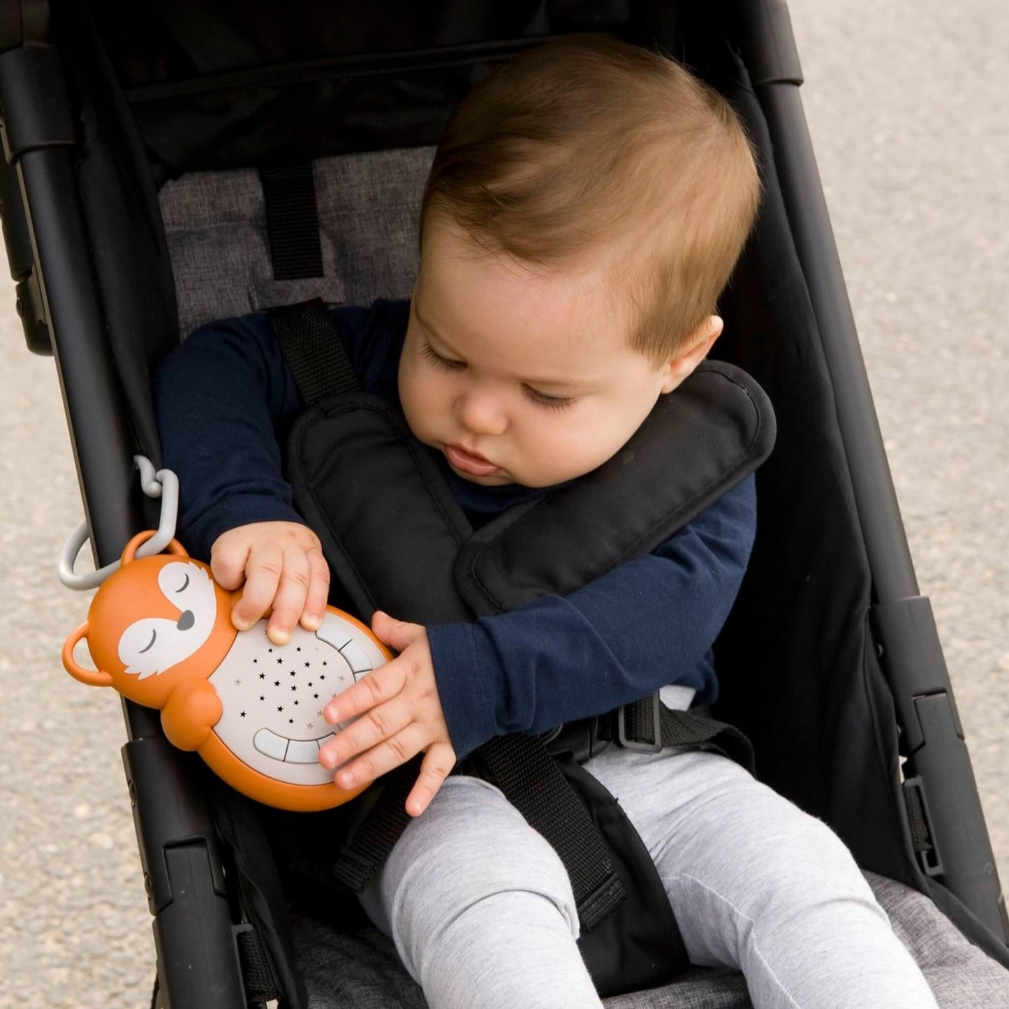 A baby seated in a pushchair exploring the fox-shaped sleep aid, showing how compact and easy to hold the portable design is.