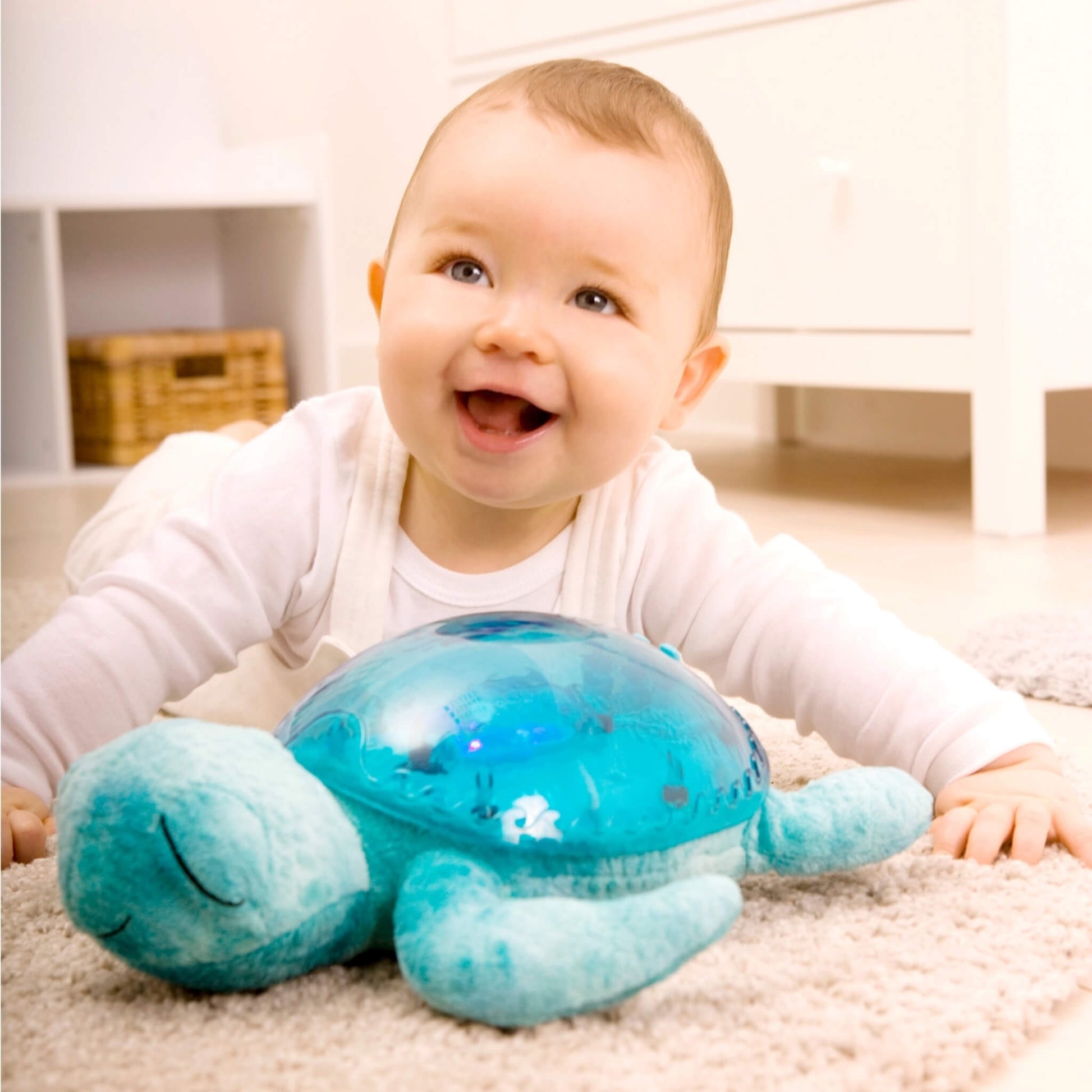 A smiling baby lying on a cream carpet beside a turquoise Tranquil Turtle, with the turtle's large translucent aqua shell in front of them.