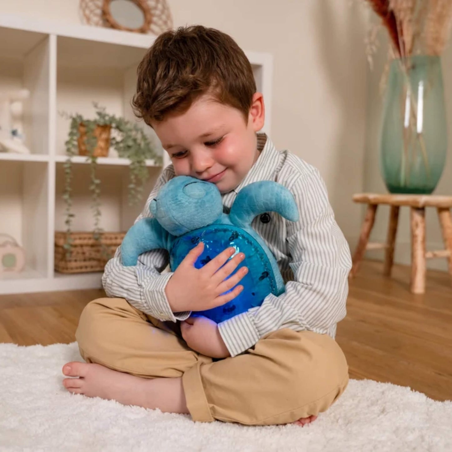 A young child sitting on the floor and cuddling a soft turquoise Tranquil Turtle against their chest while its translucent shell glows blue.