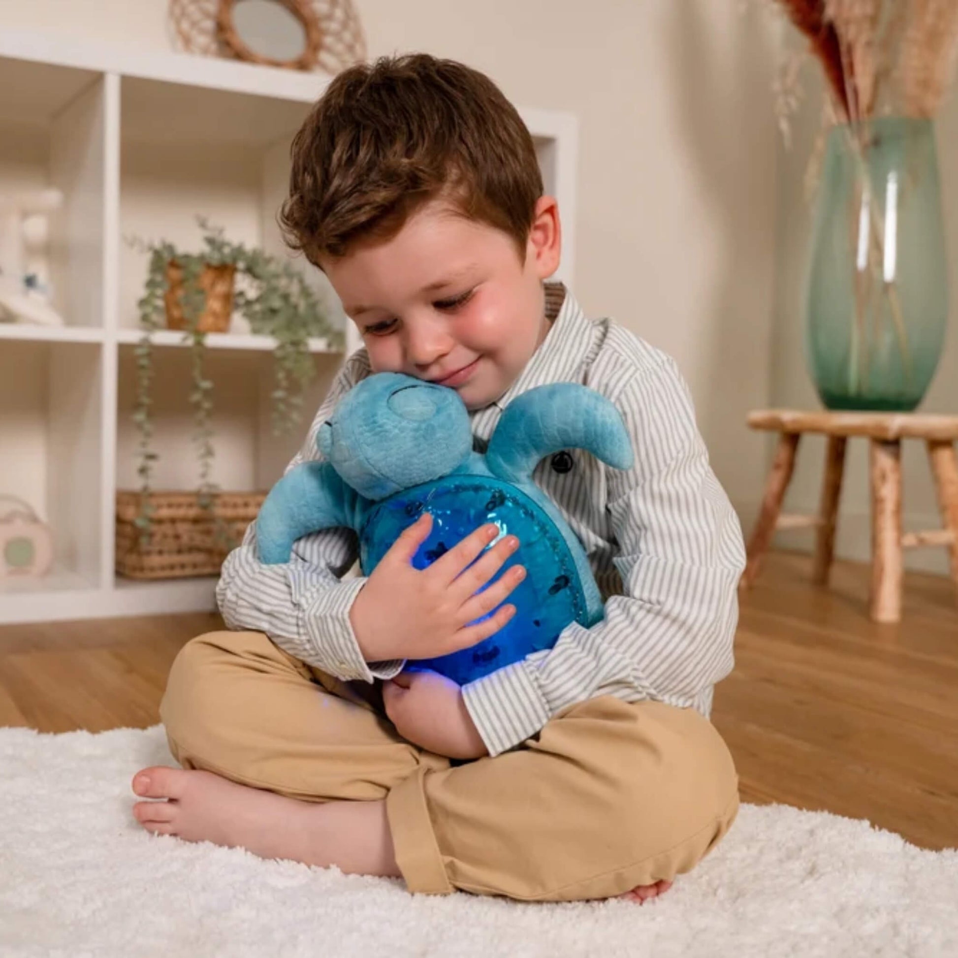 A young child sitting on the floor and cuddling a soft turquoise Tranquil Turtle against their chest while its translucent shell glows blue.