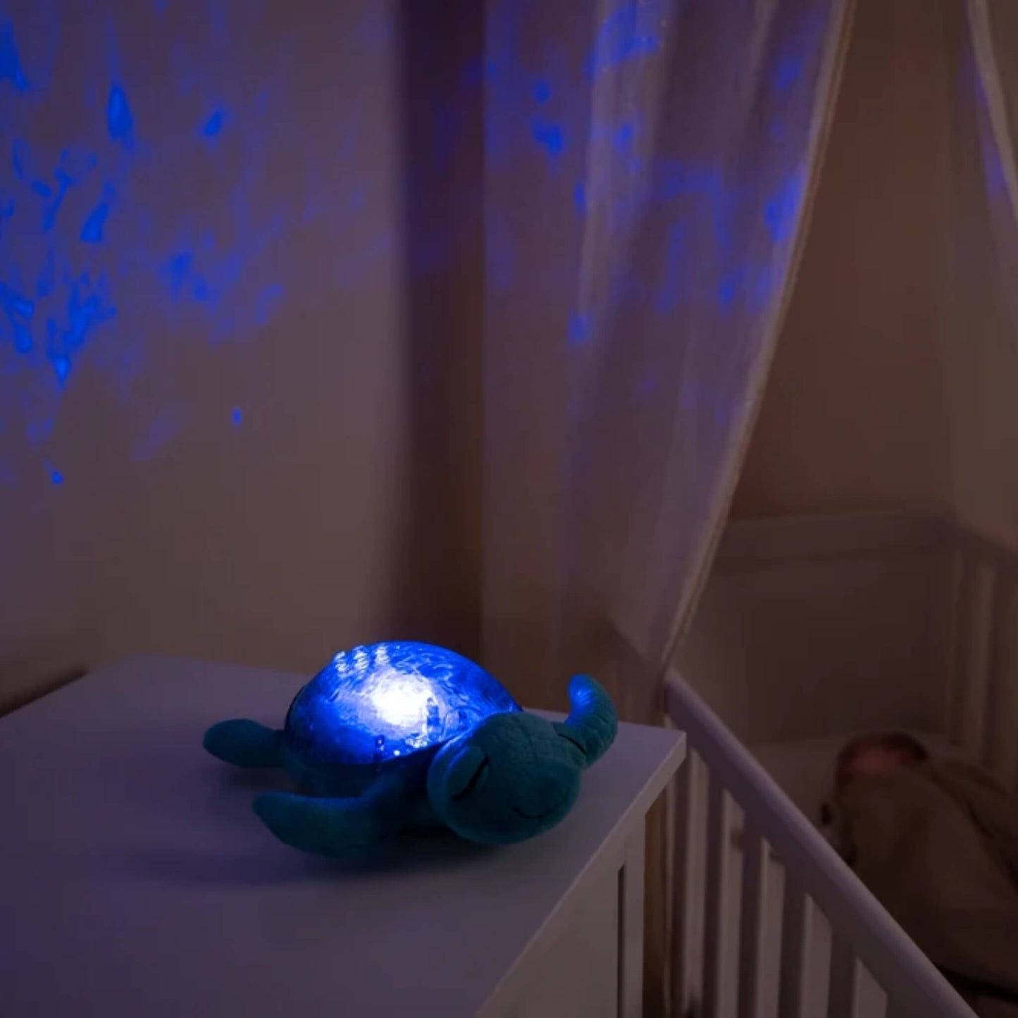 A blue sea turtle night light glowing on a white dresser in a nursery, casting a blue starry projection on the wall above a white crib.