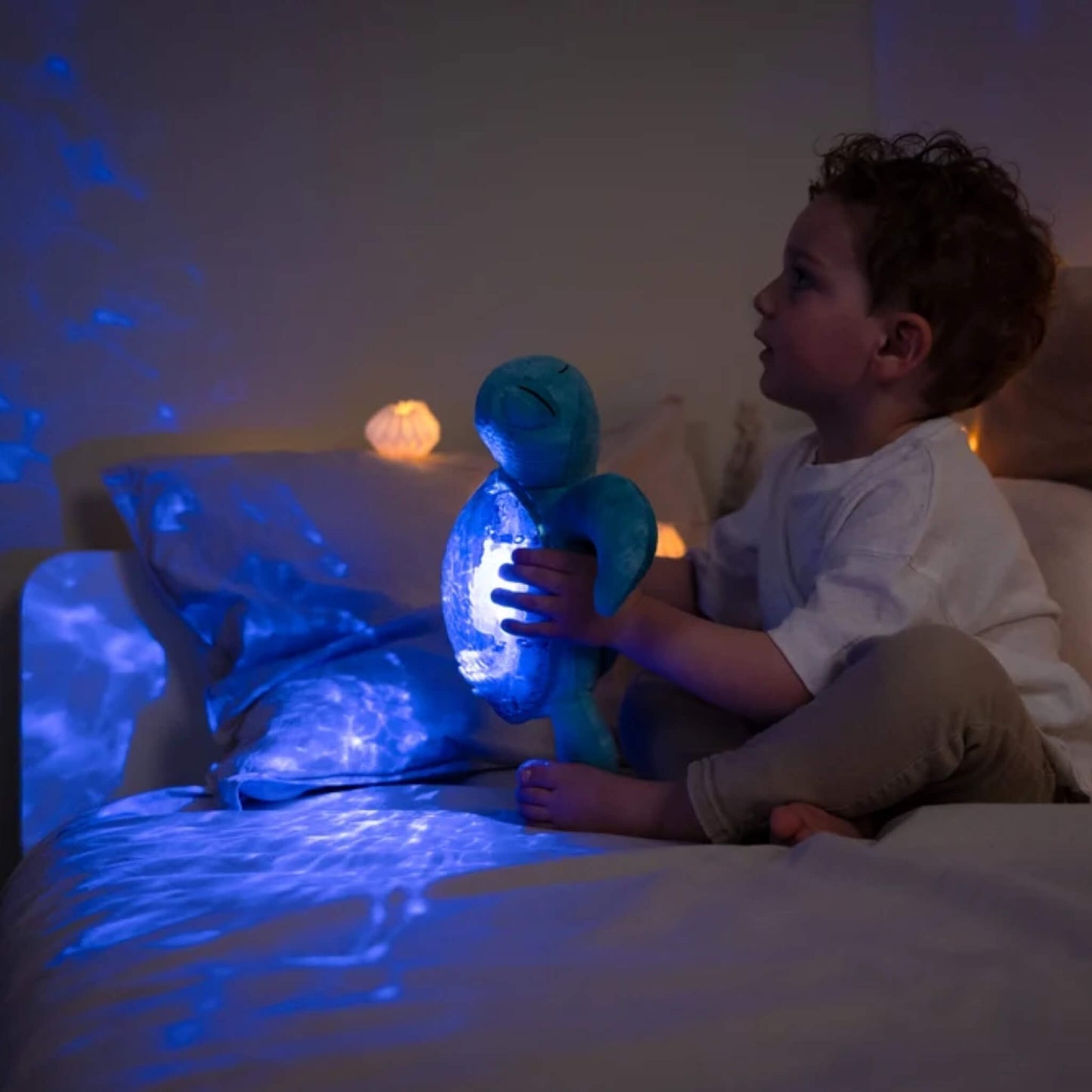 A young child sits on a bed in a dimly lit room, holding a glowing blue turtle-shaped night light that casts starry light patterns across the bedding and wall.