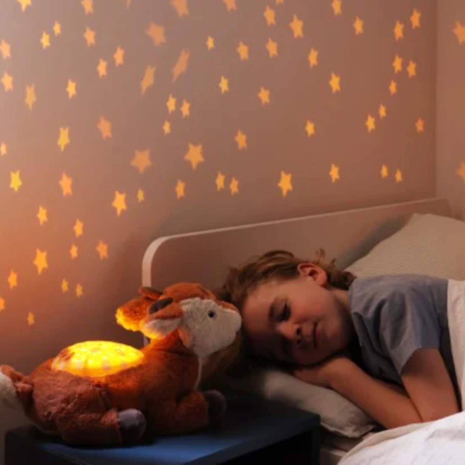A sleeping child beside a fawn night light while warm amber stars spread across the bedroom wall, showing a gentle bedside glow suitable for night-time checks.