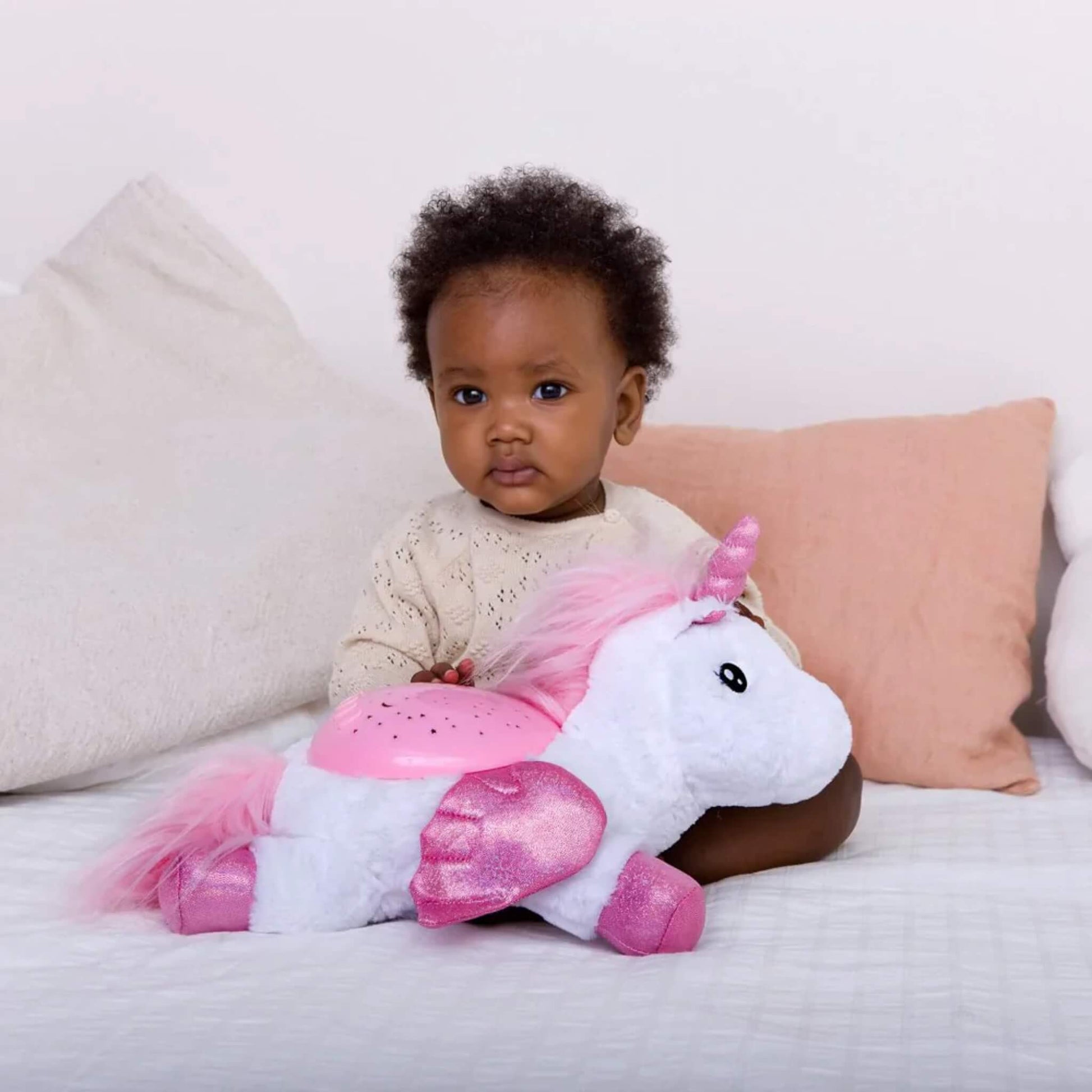 A baby sitting on a bed holding a plush unicorn night light with a pink star-projector dome and glitter wings, showing nursery-friendly size.