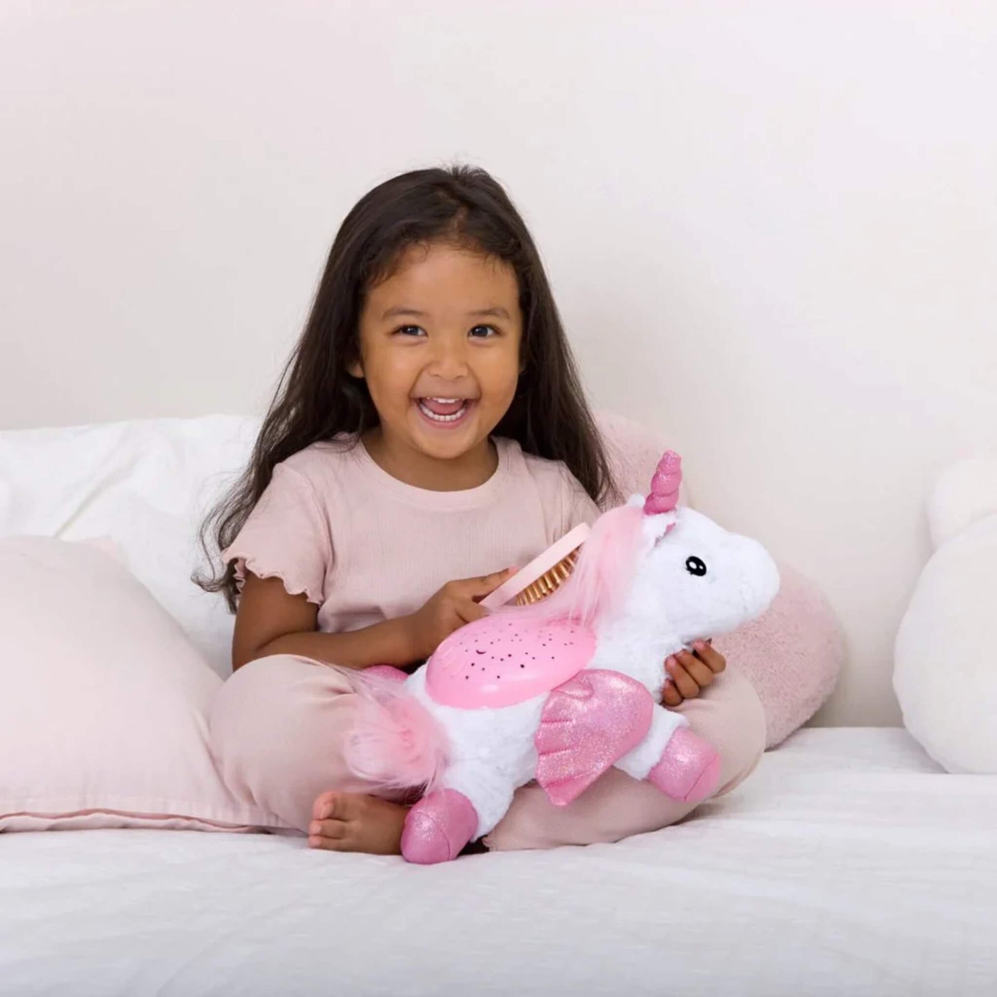 A young child on a bed brushing a plush unicorn night light’s pink mane, showing soft texture and friendly design.