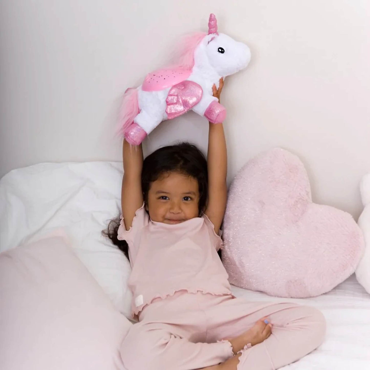 A child lifting a plush unicorn night light above their head on a bed, showing lightweight design, pink glitter hooves and a fluffy tail.
