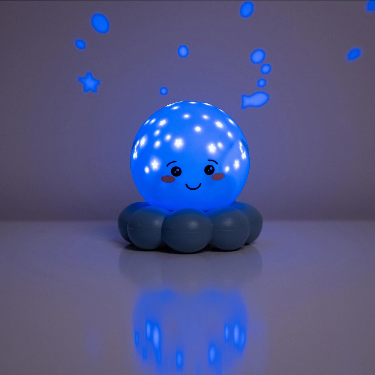 A blue octopus-shaped night light with a smiling face, sitting on a reflective surface and casting a blue star and bubble projection on a gray background.