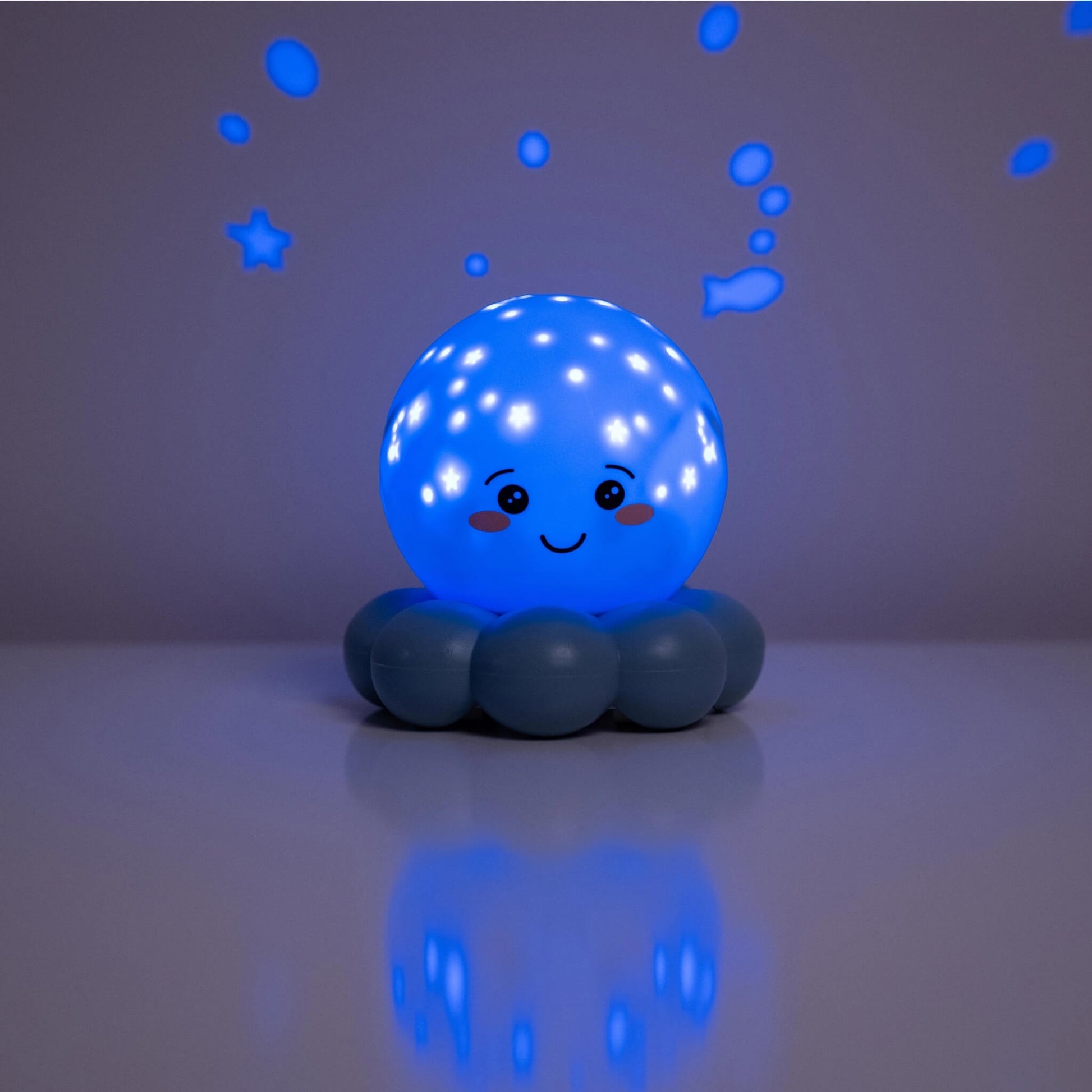 A blue octopus-shaped night light with a smiling face, sitting on a reflective surface and casting a blue star and bubble projection on a gray background.
