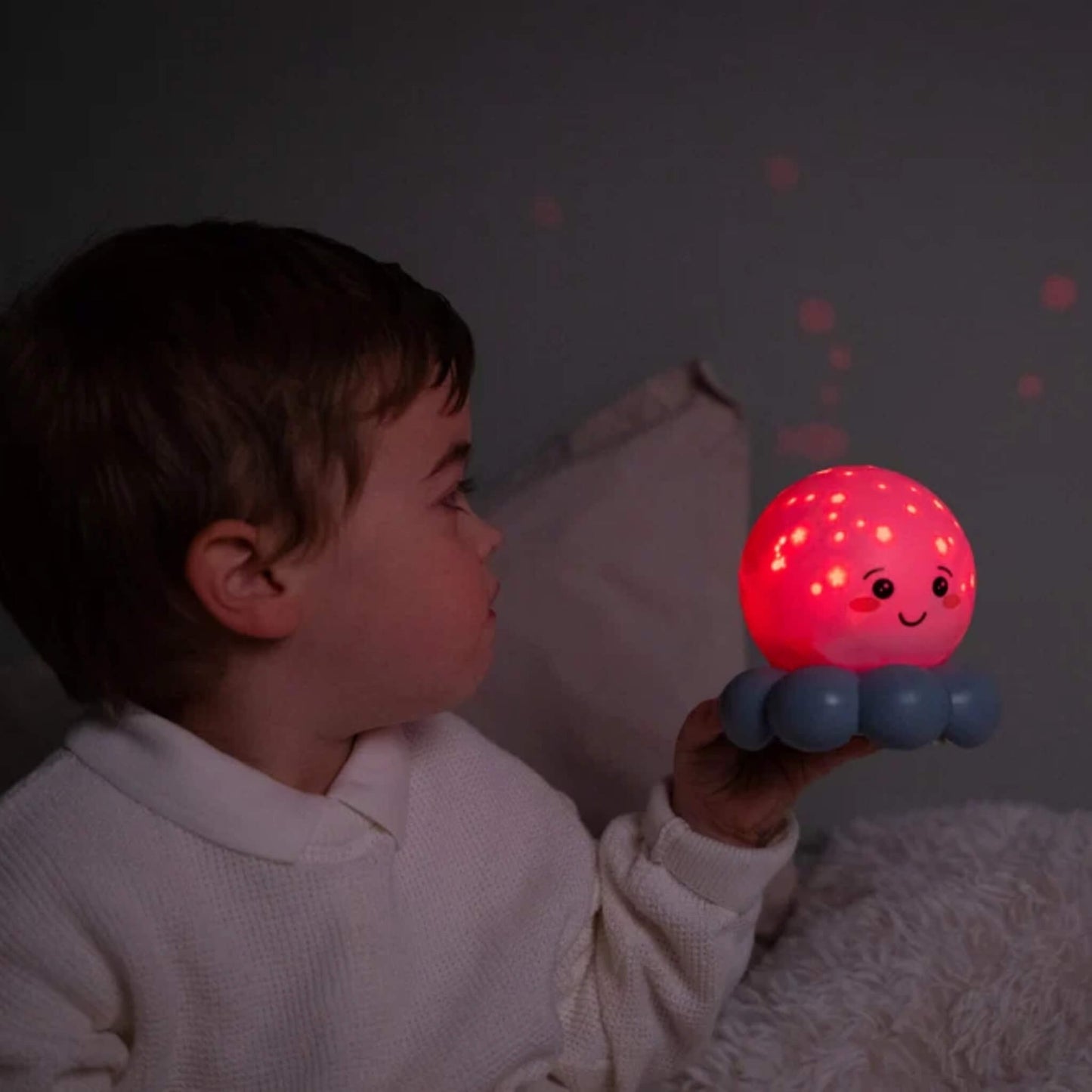 A child in a white knit top holds a small octopus-shaped night light that glows pink and red with star-like dots, while the child looks at it in a dimly lit room.