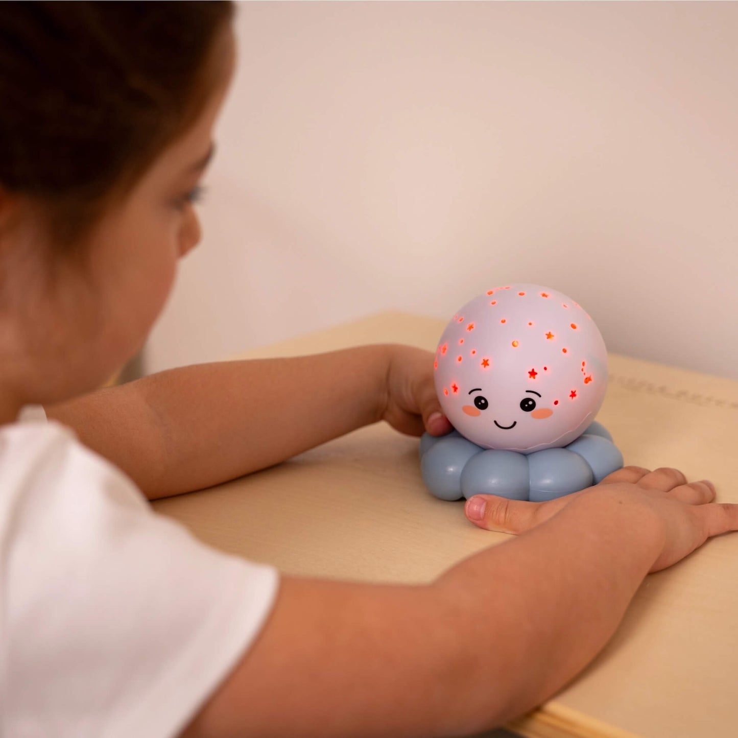 A child sitting at a desk touching a small octopus-shaped night light that projects orange stars onto a light purple surface.