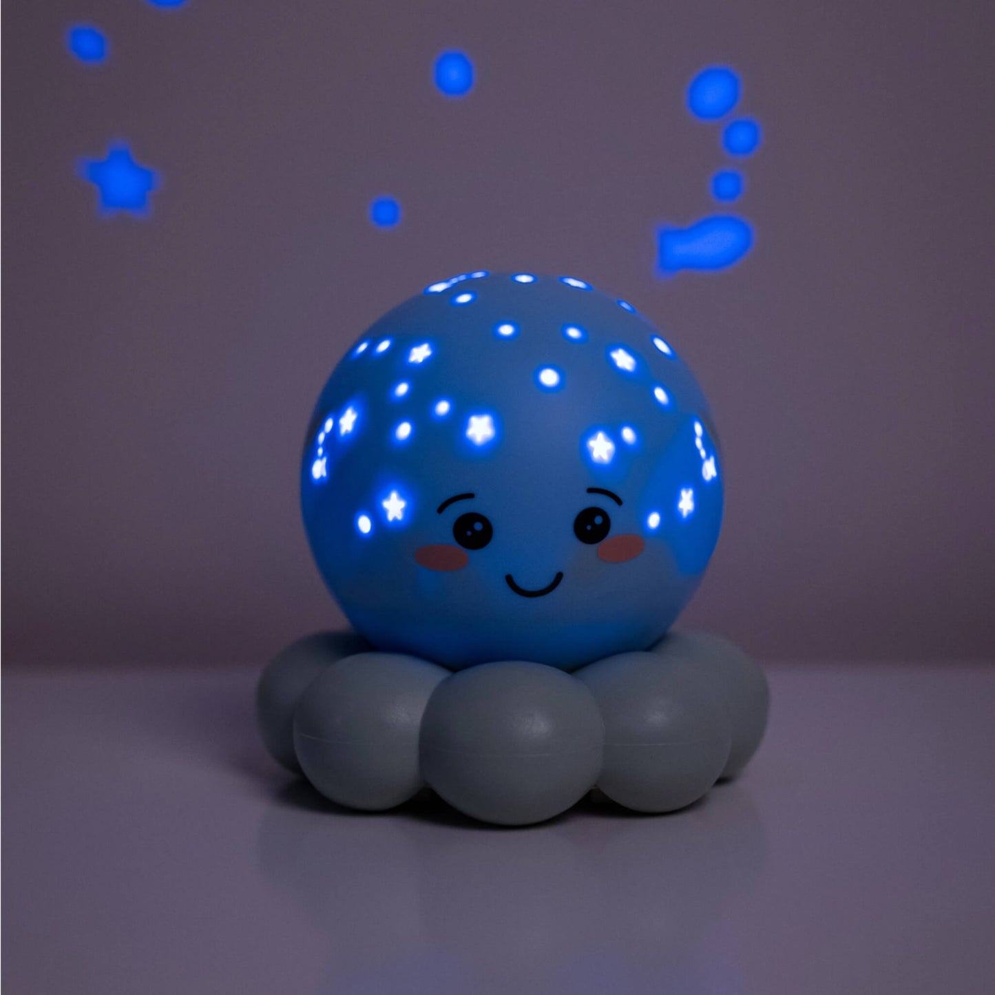 A cute octopus-shaped night light with a smiling face, casting glowing blue star and bubble projections on a dark wall and floor.