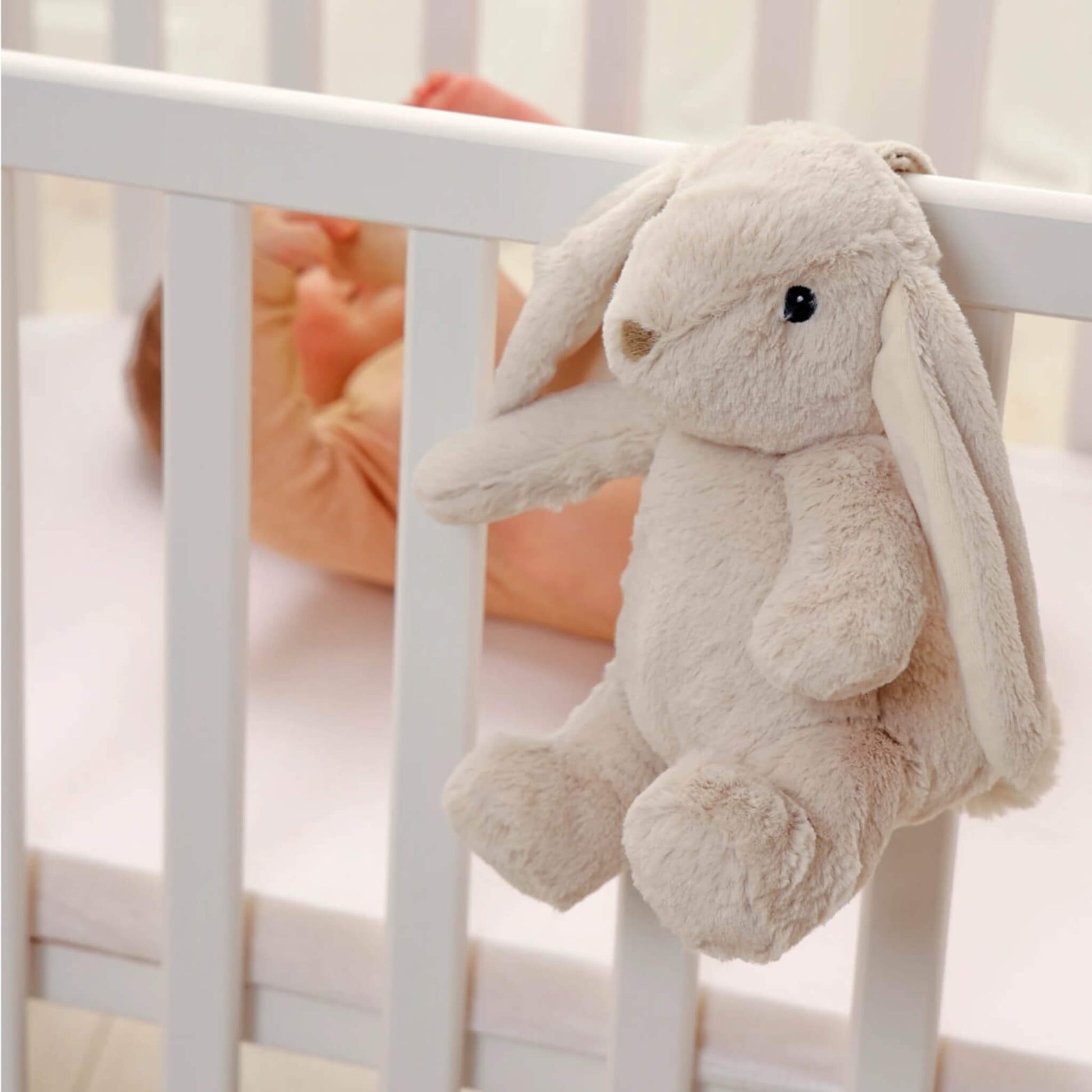 Soft bunny toy hanging by its strap from the side of a white cot, with a baby lying inside.