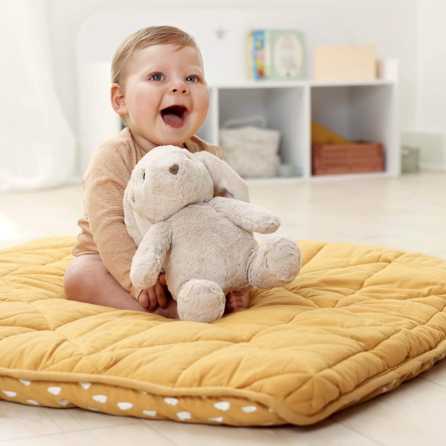 Smiling baby sits on a quilted yellow mat, holding a plush bunny toy with floppy ears.